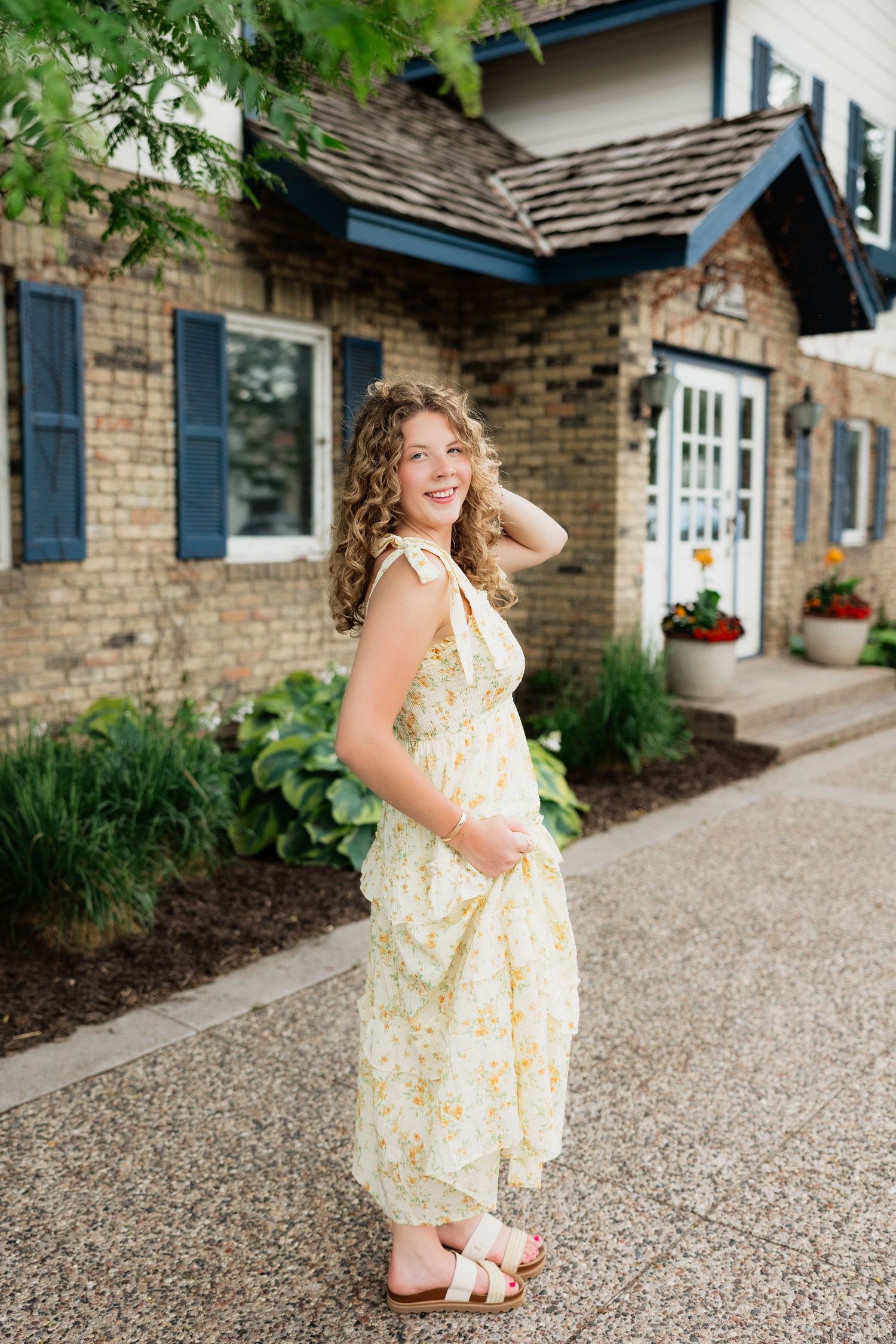 Young woman in yellow floral midi dress smiling outdoors in front of brick building with blue shutters.