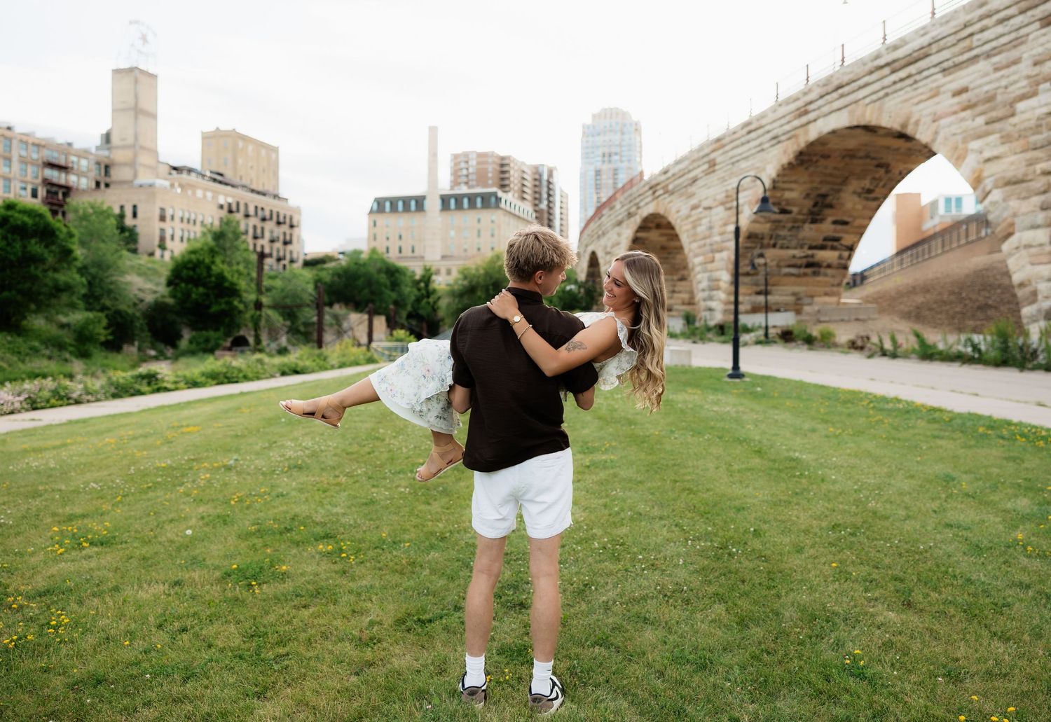 Couple embracing on green grass near Stone Arch Bridge in Minneapolis with city skyline in background.
