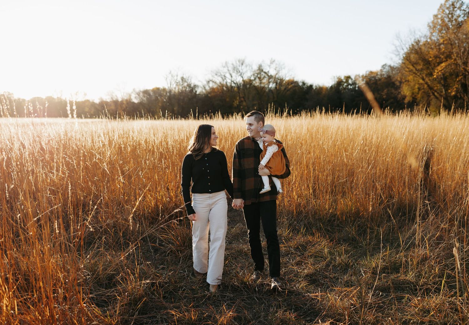 Young couple walking hand in hand through golden autumn field while carrying their baby girl.