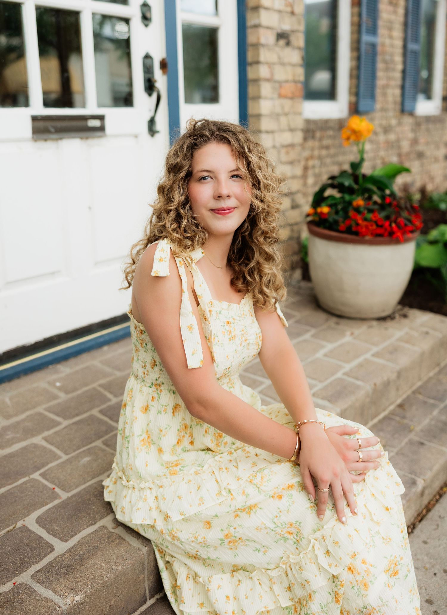 Young woman with curly hair sits on steps in a floral yellow sundress beside a white door and flower pot.