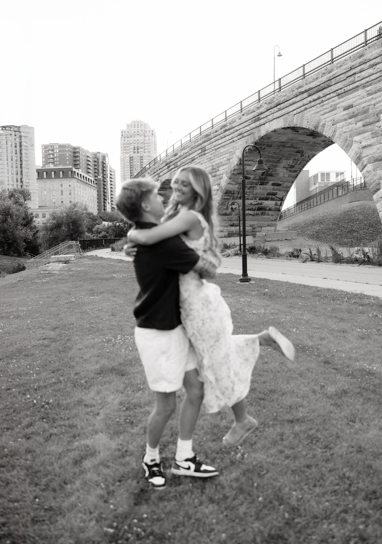 Black and white photo of couple embracing and kissing near stone arch bridge on grassy riverbank.