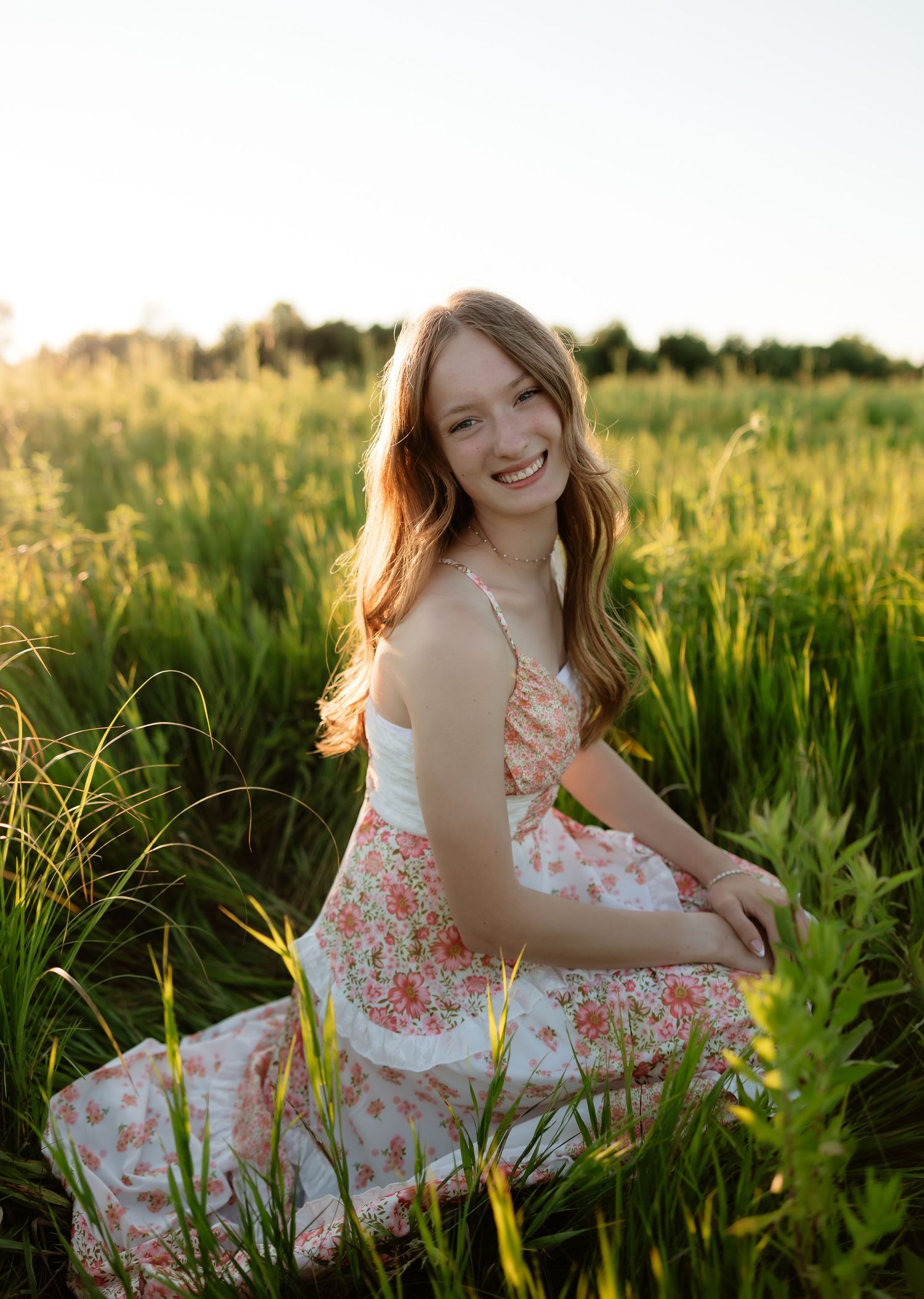 Young woman in floral dress sitting in green field, smiling warmly in golden hour sunlight.