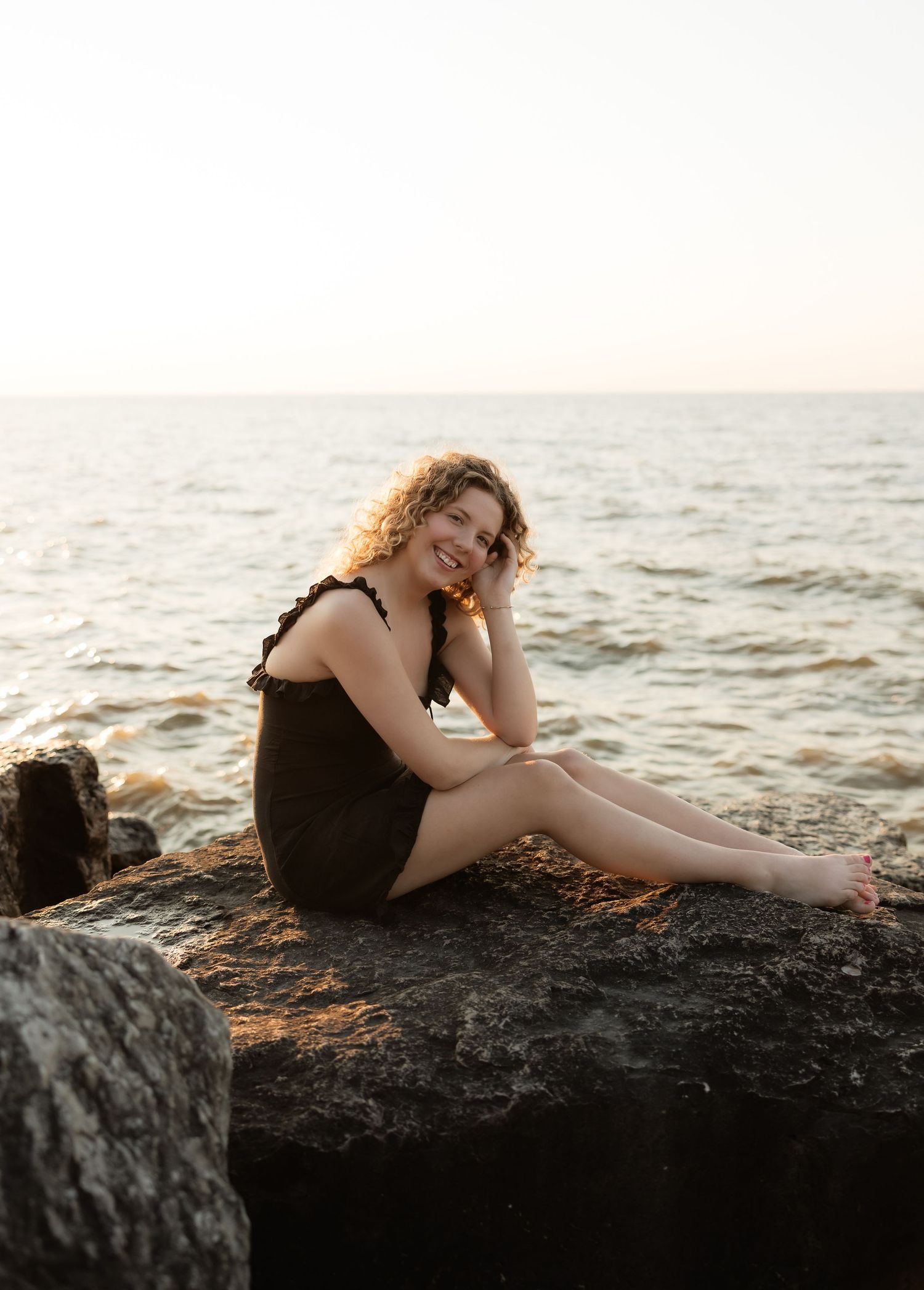Cheerful woman with curly hair wearing black dress posed barefoot on coastal rocks at sunset.