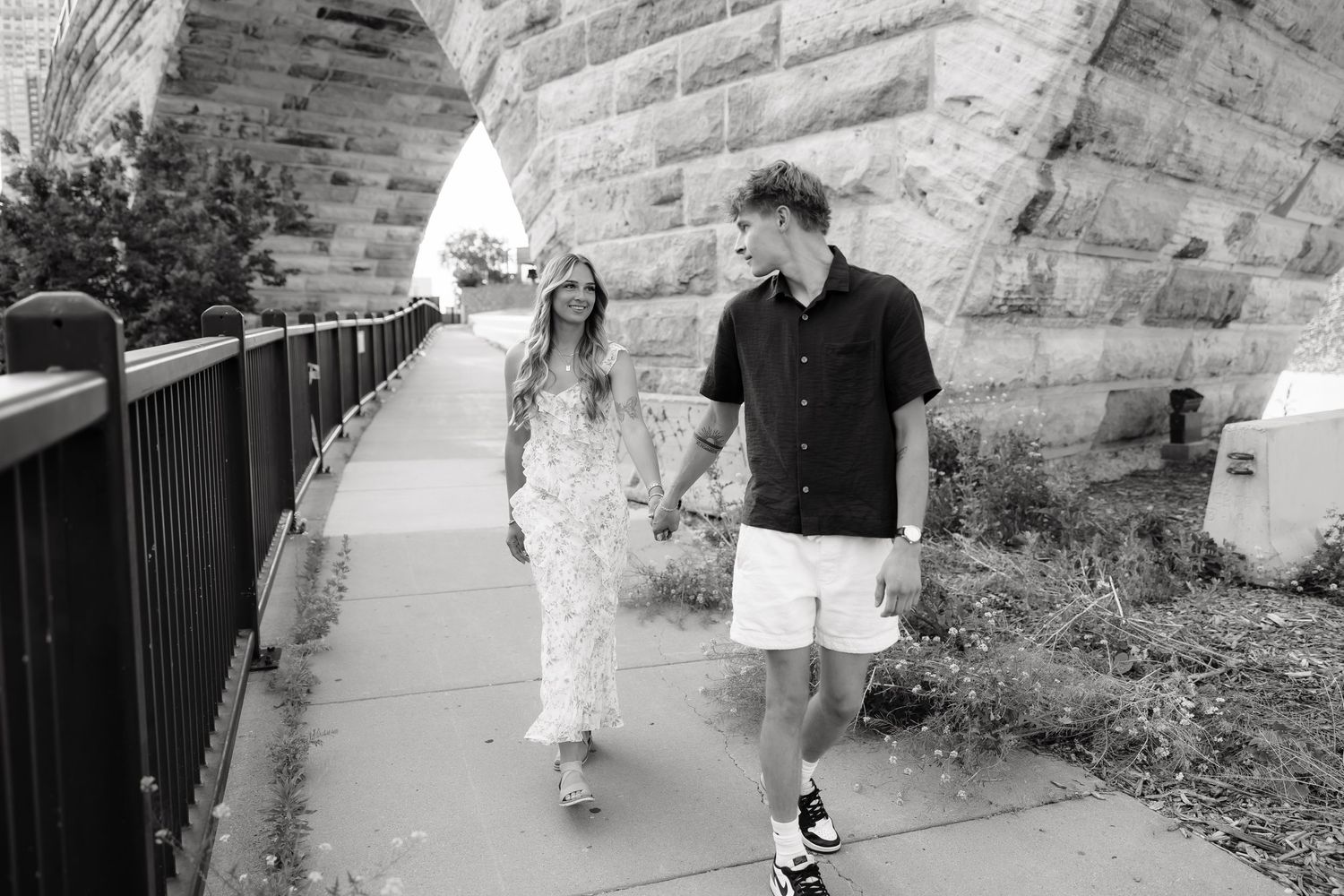 Couple holding hands walking along a stone arch bridge pathway in a black and white romantic outdoor photo.