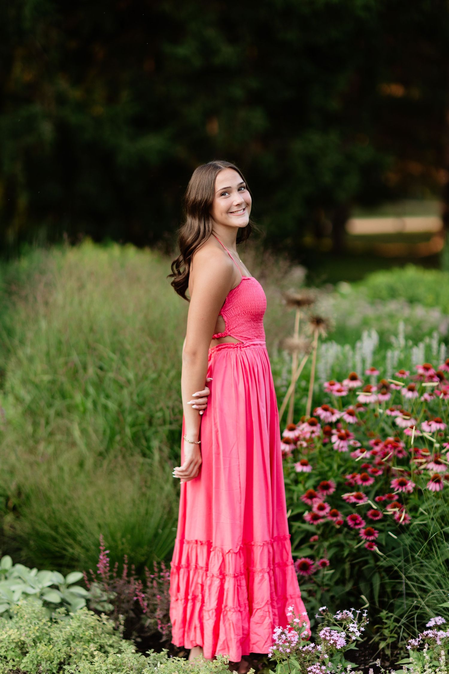 Smiling teen girl in pink spaghetti strap maxi dress standing among colorful garden flowers outdoors.