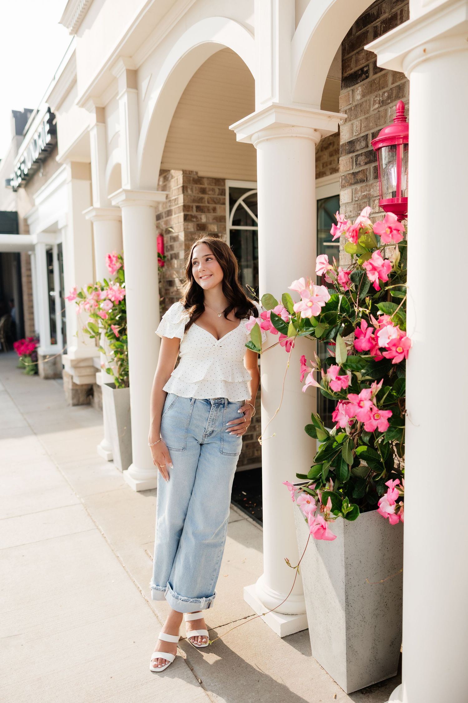 Young woman in white top and jeans posing by white columns with pink flowers at an outdoor shopping center.