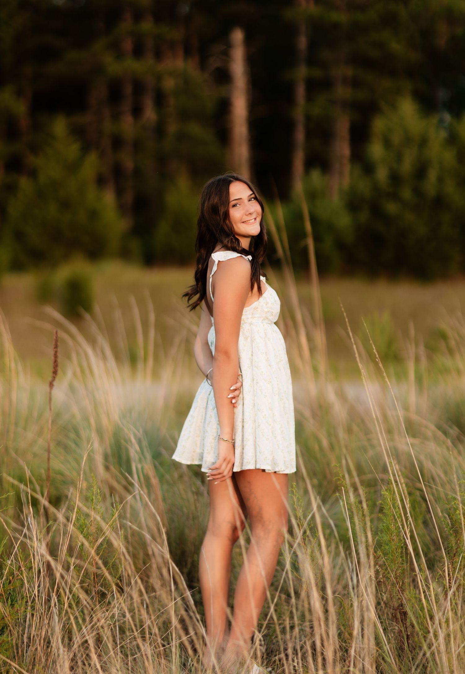 Brunette teen in white sundress standing in tall golden grass with pine trees in background.