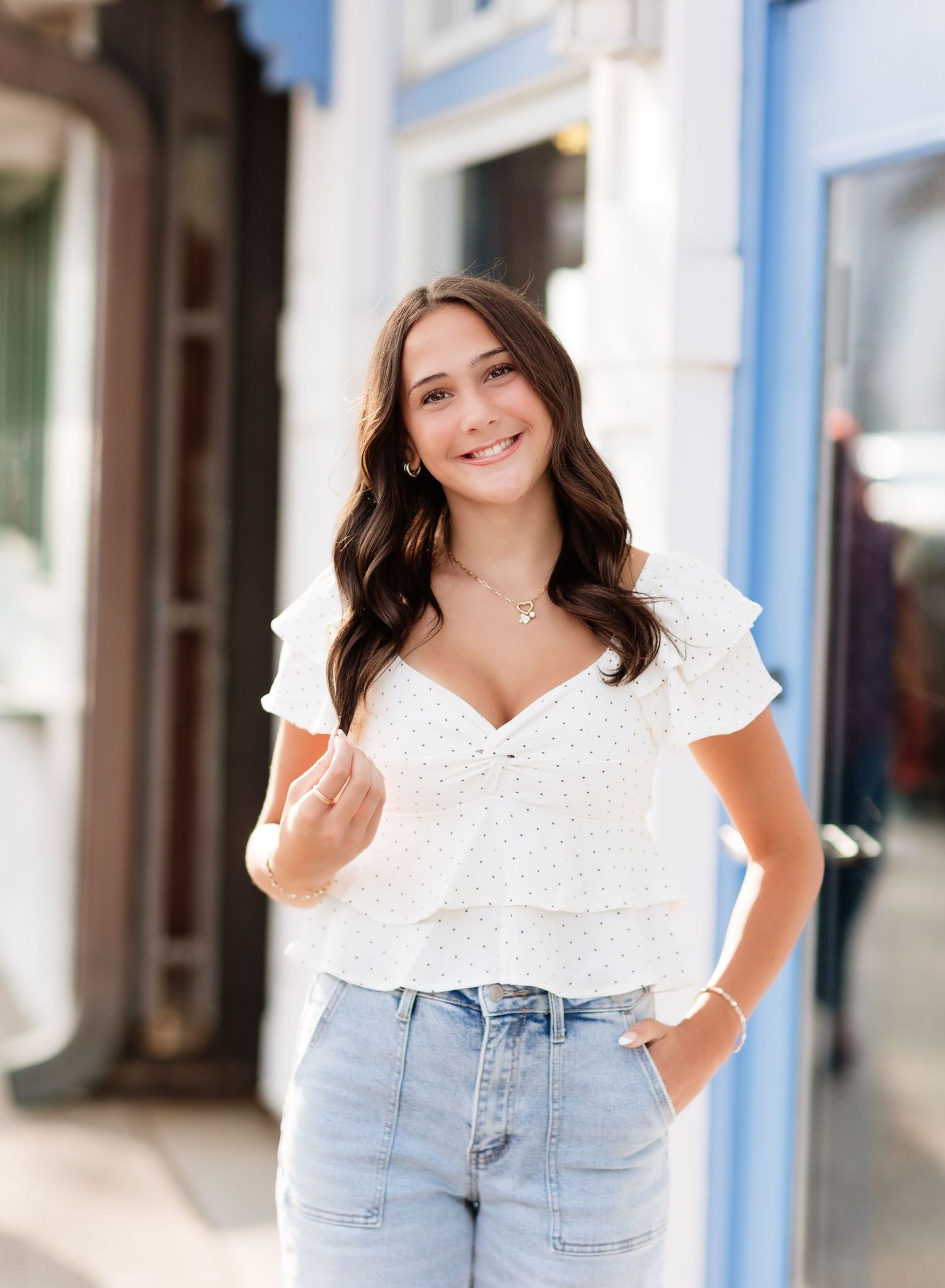 Smiling brunette woman in white ruffled top and light jeans posing outdoors on a sunny day.