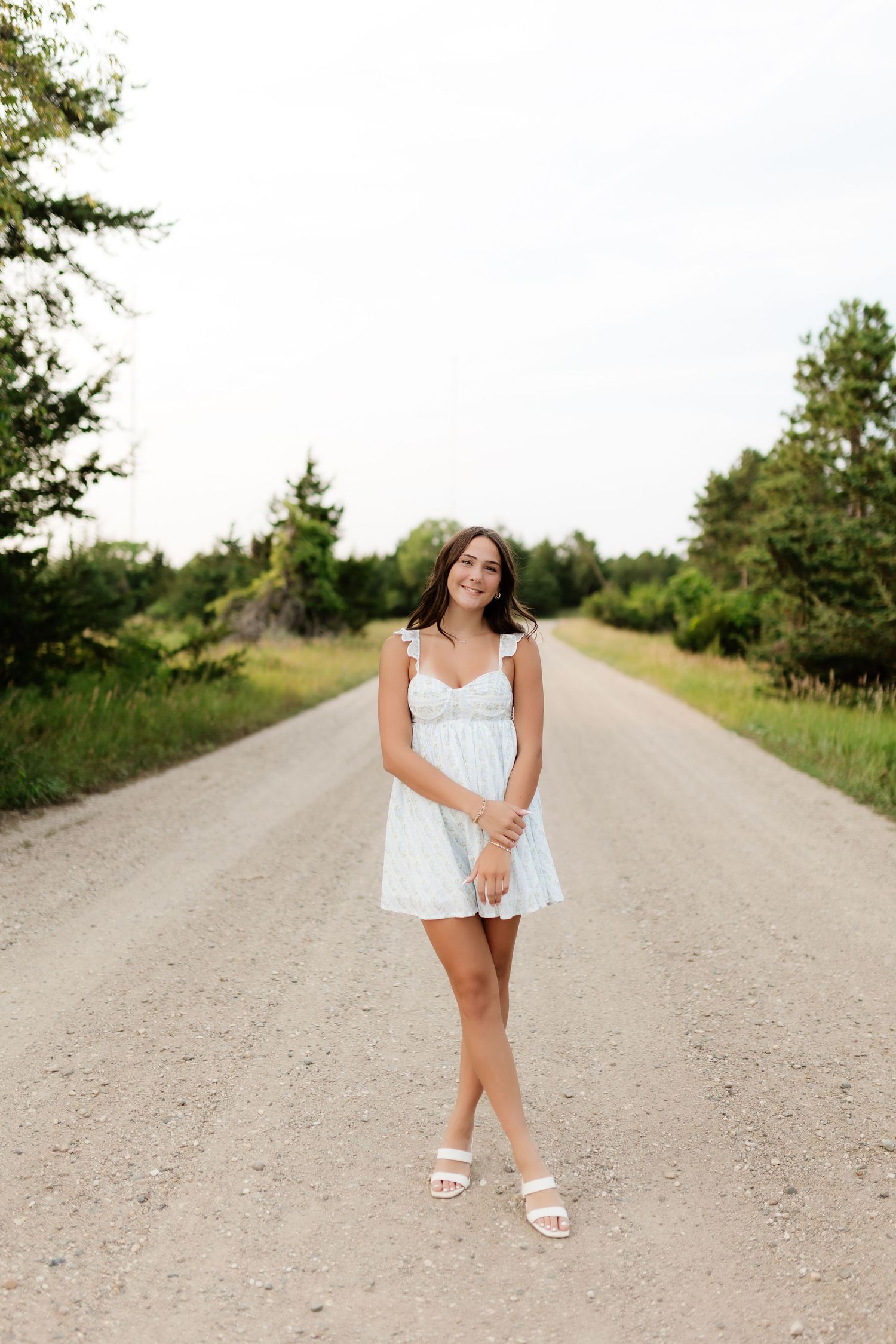 Smiling young woman in white mini dress standing on a scenic gravel road lined with green trees.