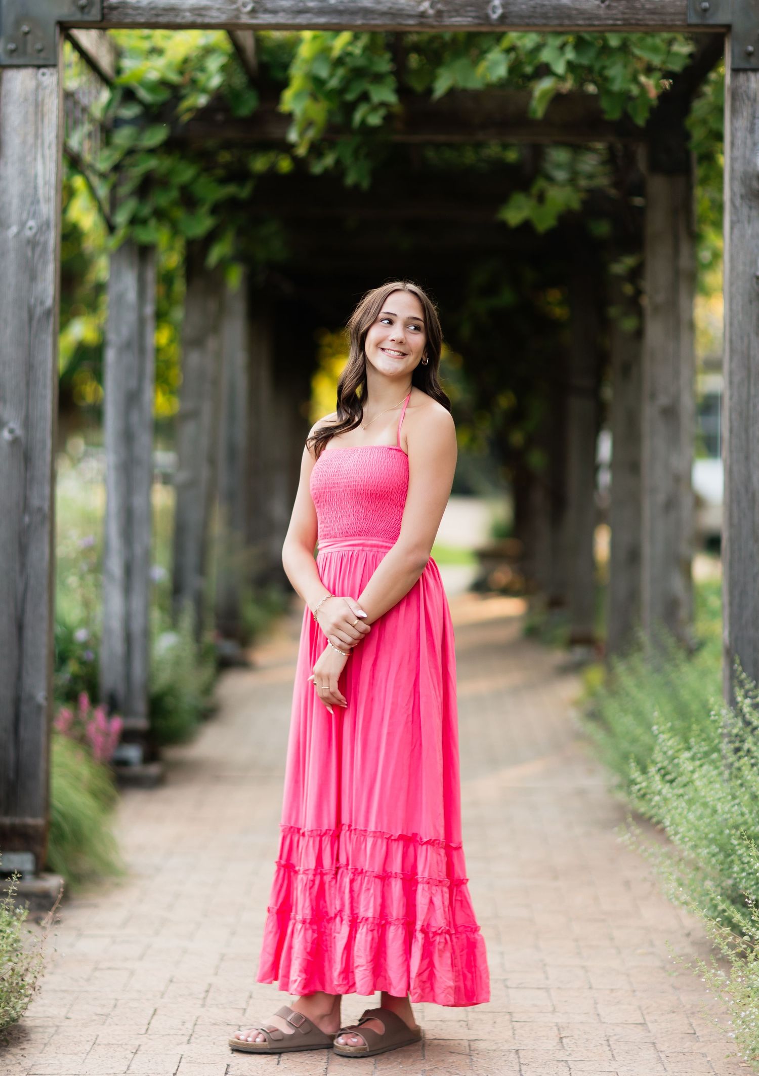 Young woman in pink ruffle maxi dress standing under wooden pergola archway in lush garden setting.