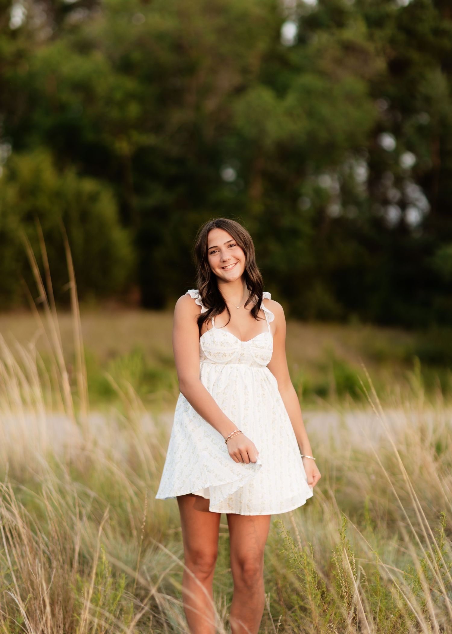 Smiling brunette in white sundress posing in tall grass with green trees behind.
