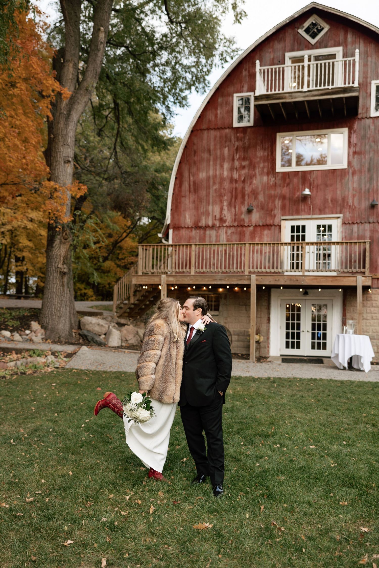 Bride and groom kissing in front of a rustic red barn surrounded by vibrant fall foliage.