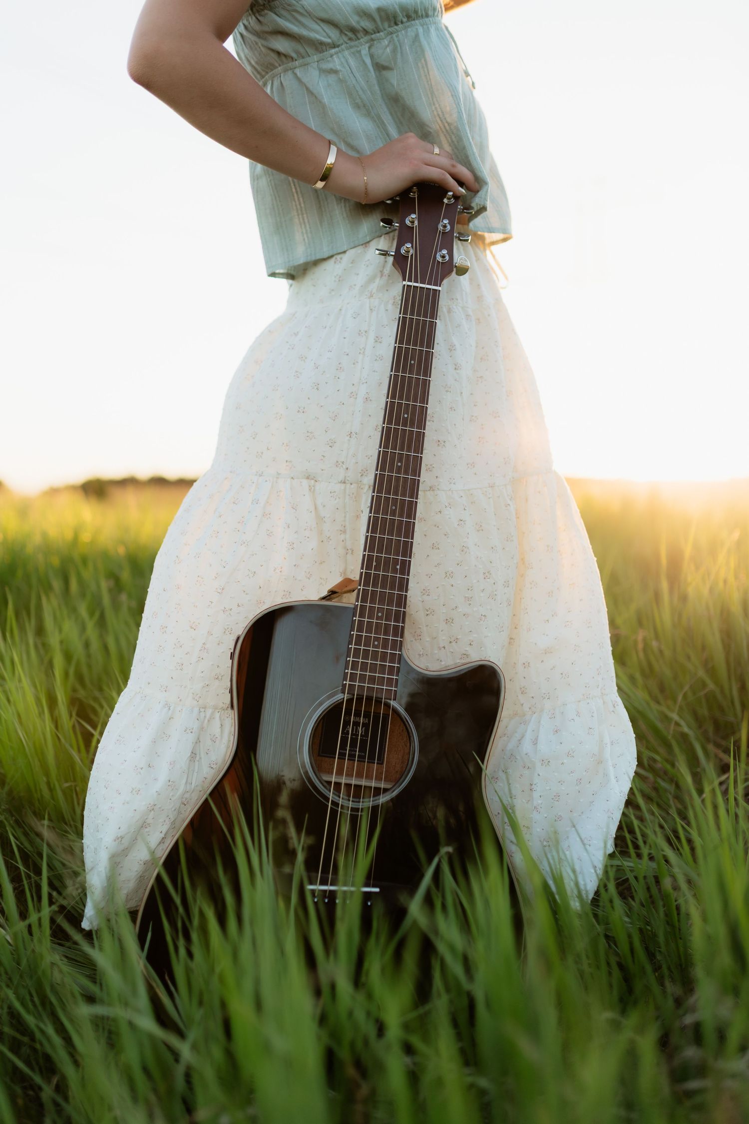 Woman in white maxi skirt standing in tall grass field holding black acoustic guitar at sunset.
