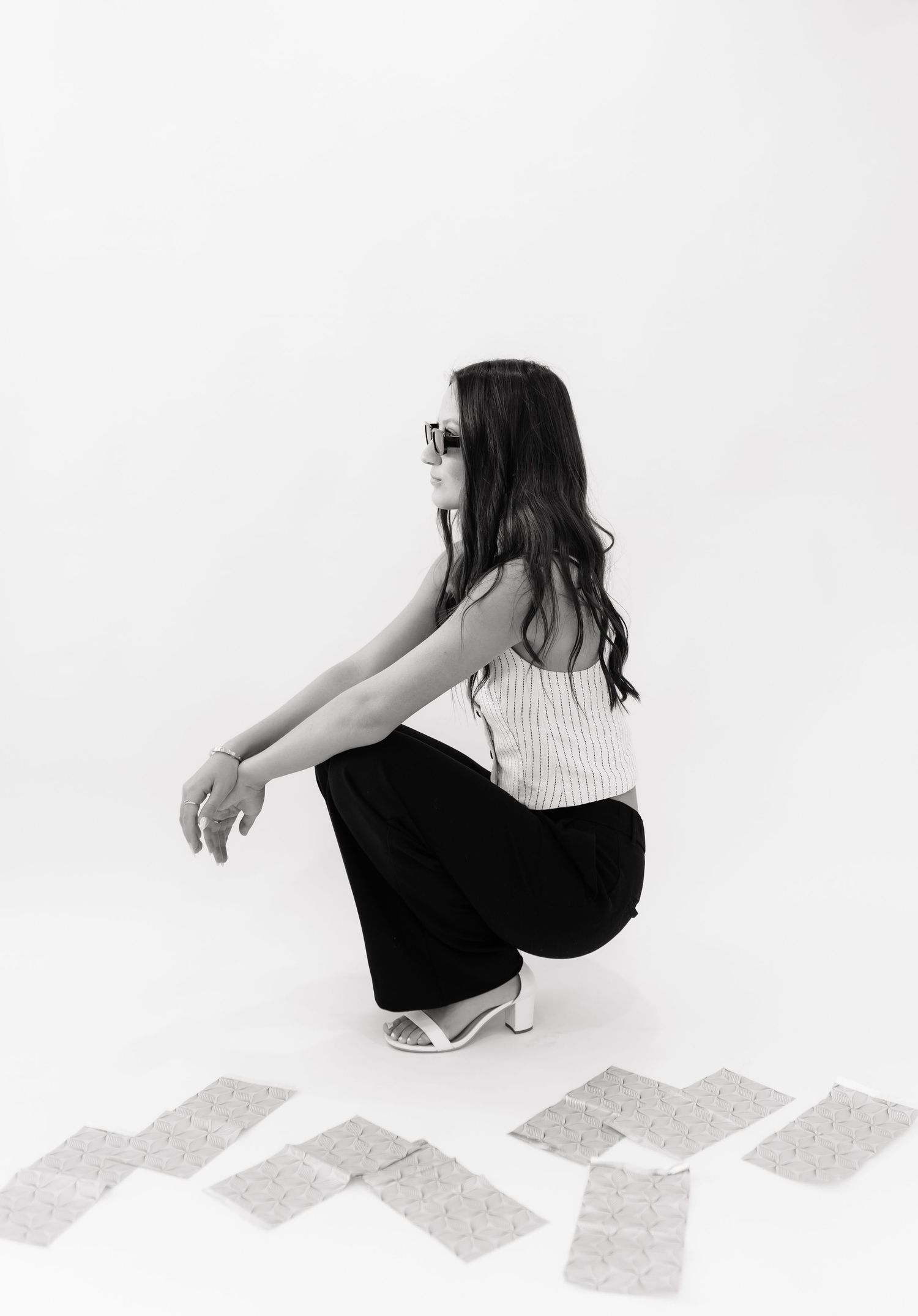 Black and white photo of brunette woman crouching in sunglasses, striped top, and wide-leg pants.