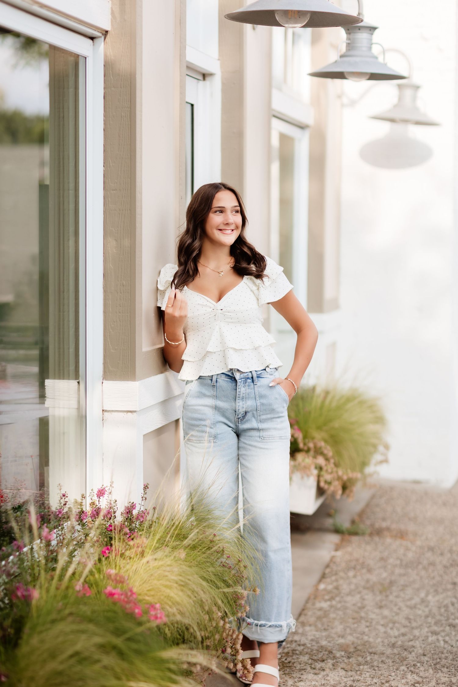 Smiling brunette in white tiered ruffle top and cuffed wide-leg jeans leaning against building outdoors.