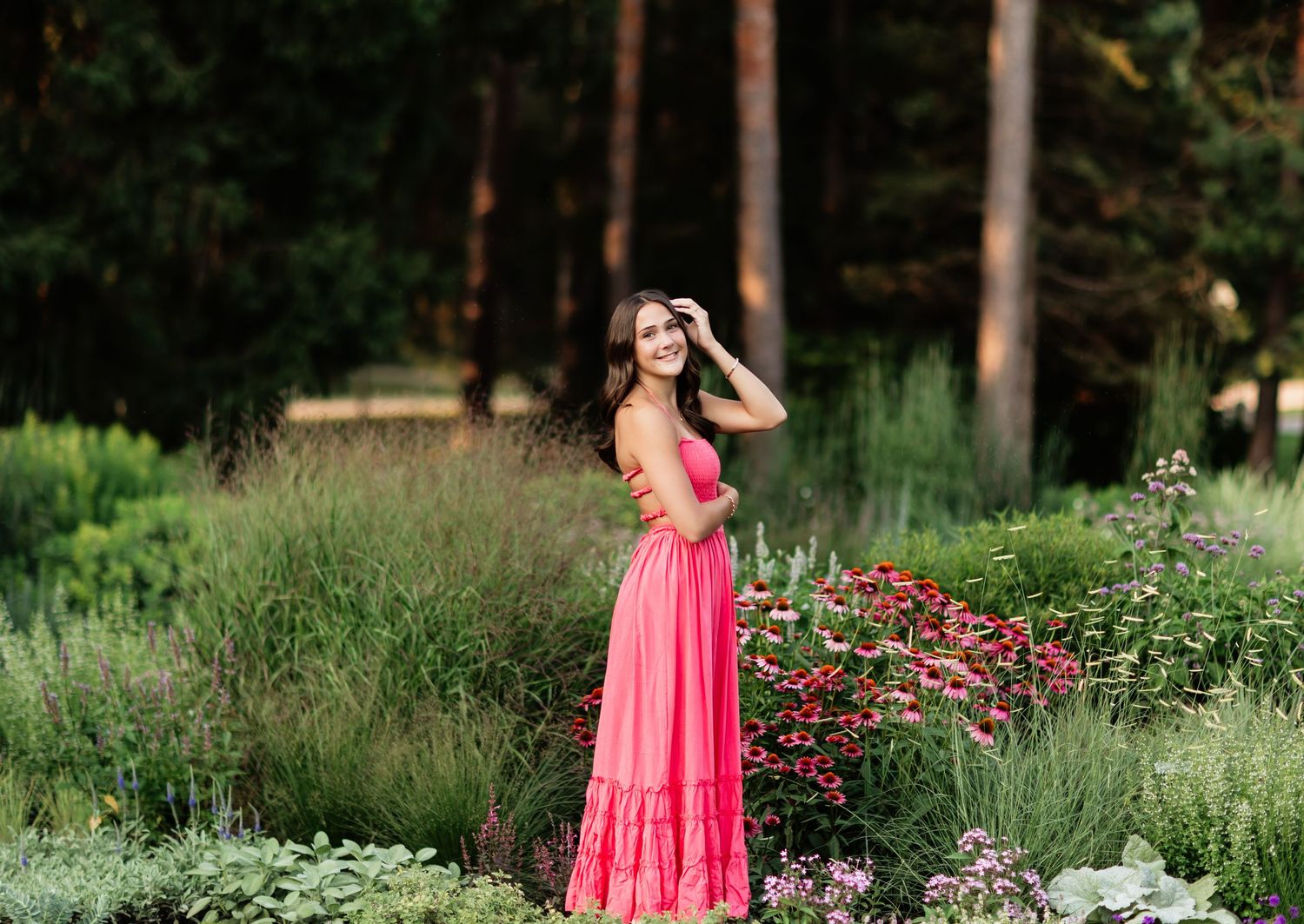 A woman in a pink maxi dress poses gracefully among colorful garden flowers with tall trees in the background.