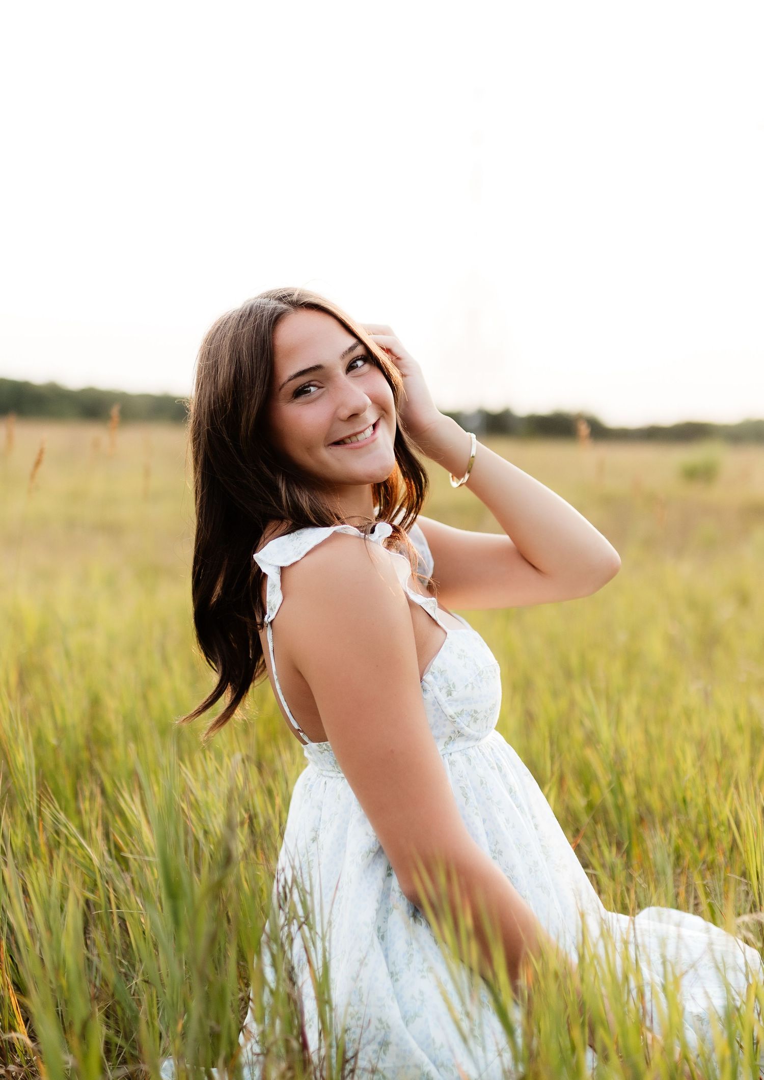 Smiling young woman in light blue dress sitting in tall green grass at golden hour.