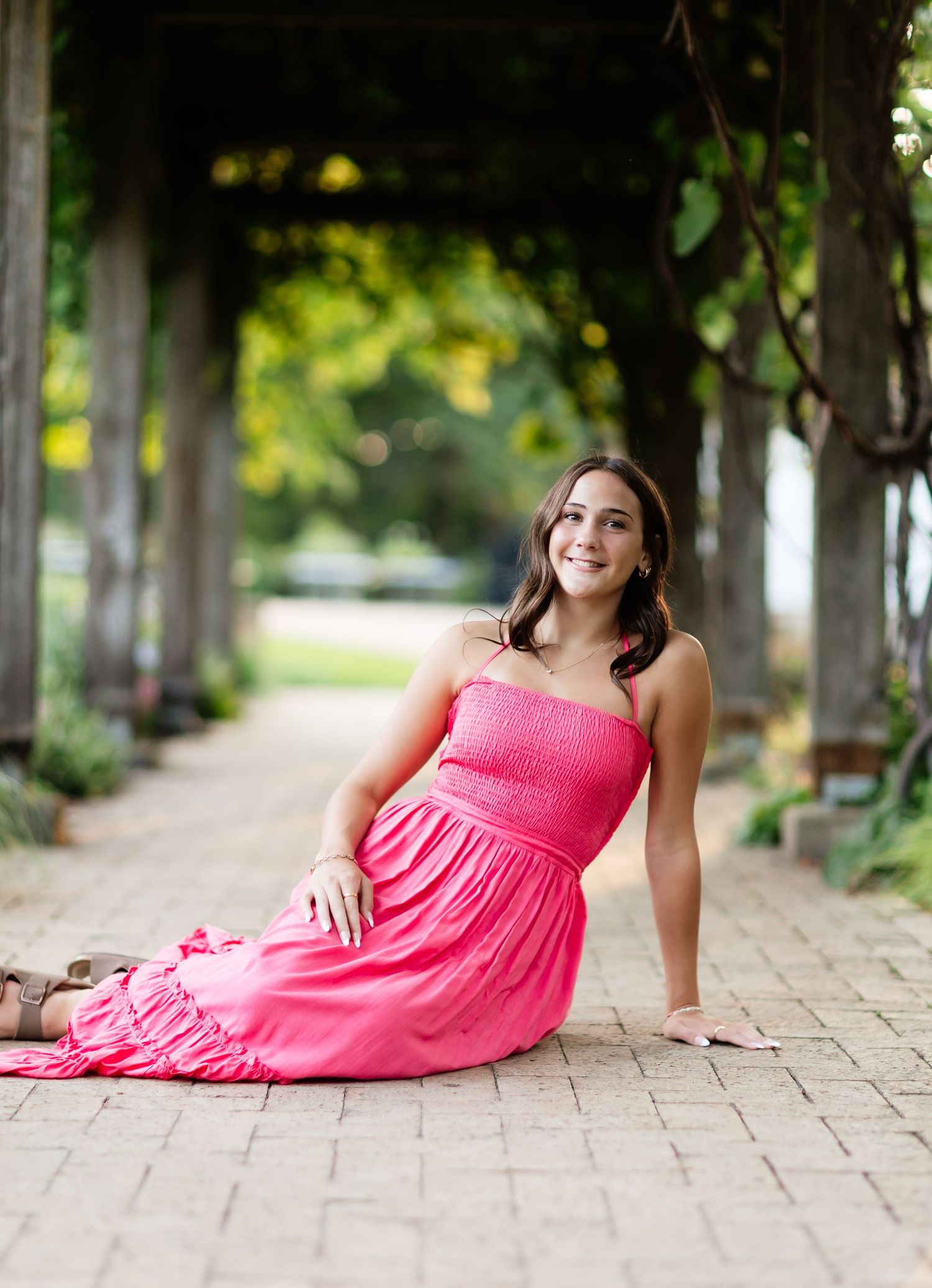 Young woman in bright pink dress sitting gracefully on brick path under green tree canopy.
