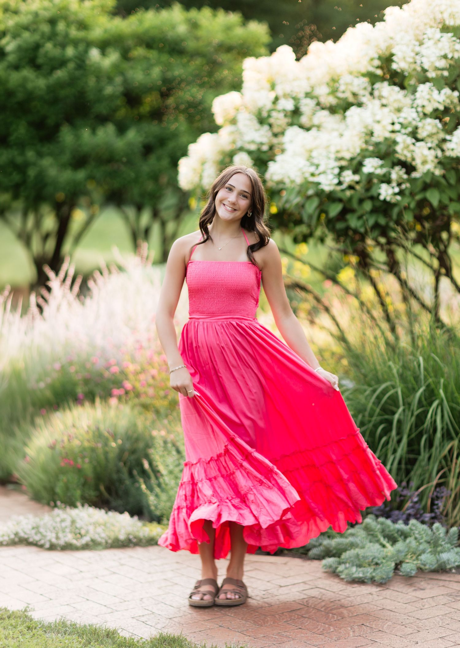 Young woman in bright pink maxi dress twirling in a lush garden with blooming flowers.