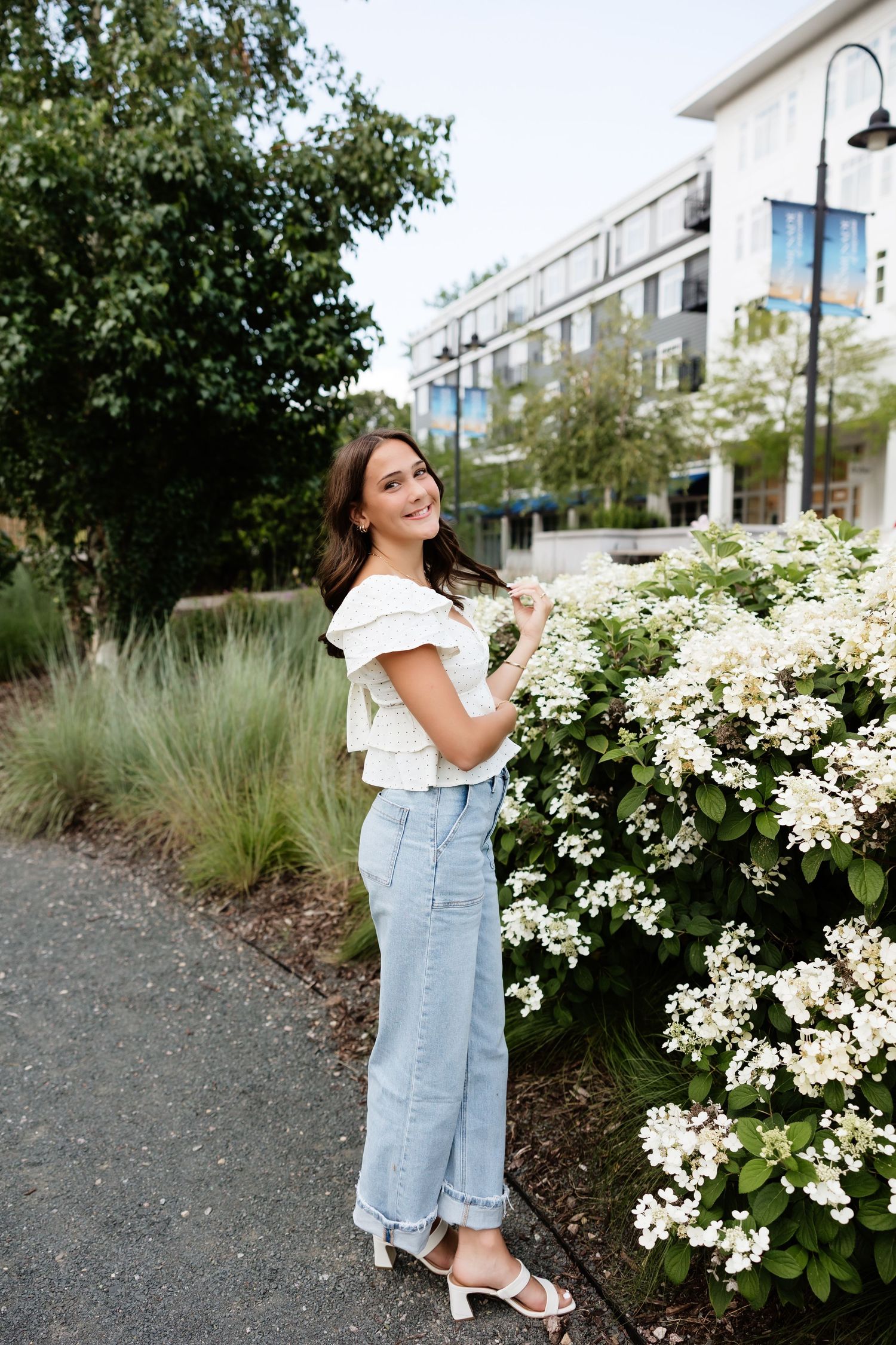Young woman in white ruffled top and light jeans smiling beside white flowering bush in urban garden.