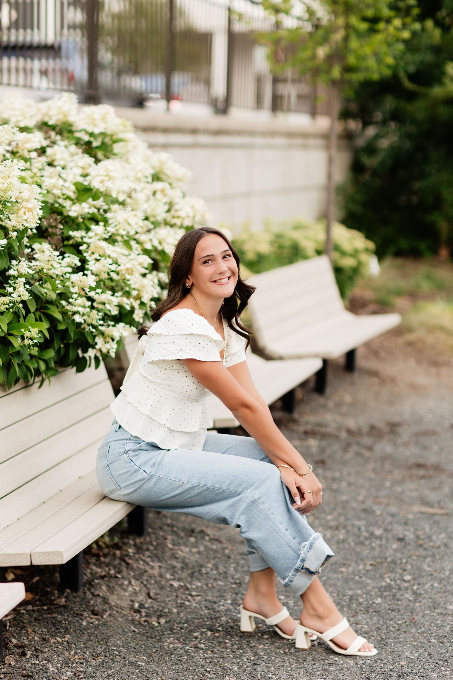 Young woman in white off-shoulder top and jeans sitting on a bench beside blooming white hydrangeas, smiling outdoors.