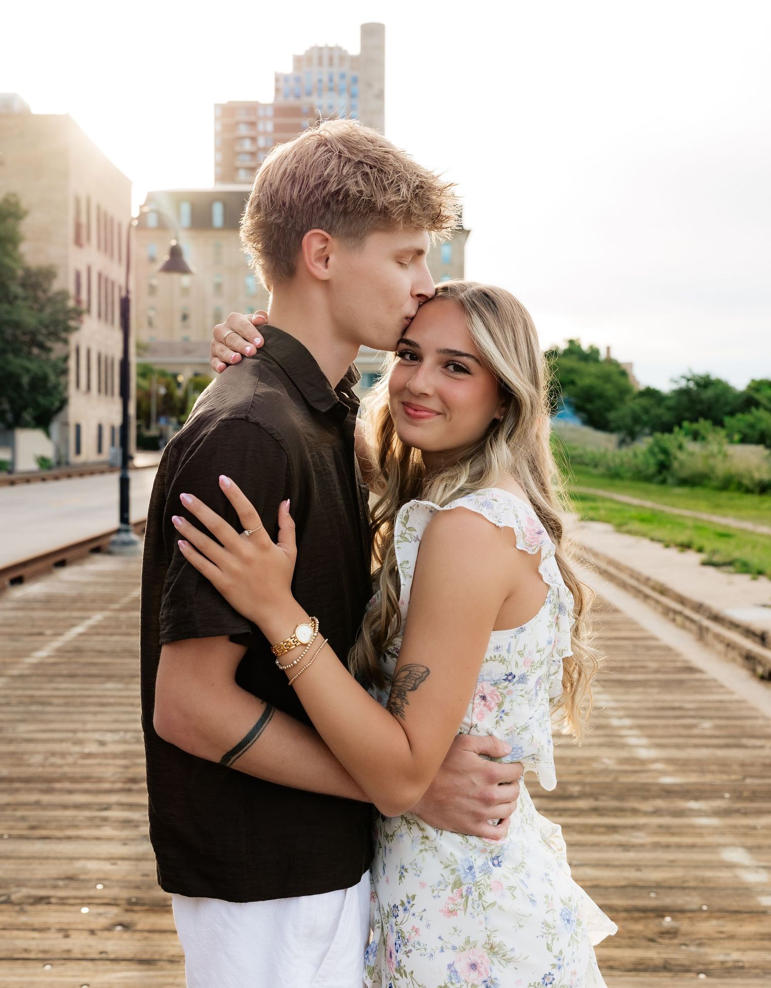 Young couple embracing on urban bridge, man kissing woman's forehead, she smiles at camera in floral dress.
