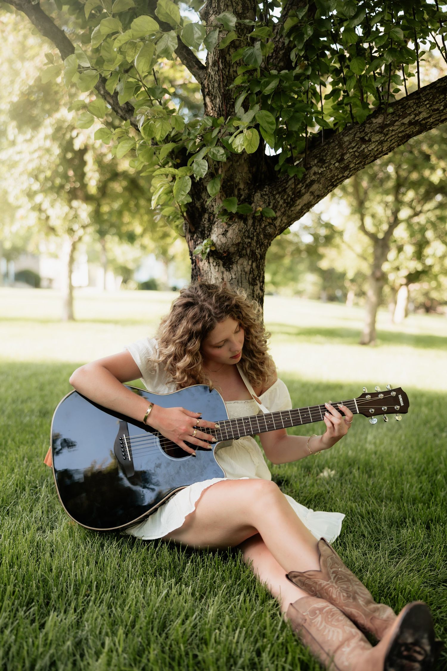 Young woman with curly hair playing acoustic guitar while sitting on green grass under a tree in a white dress and cowboy boots.