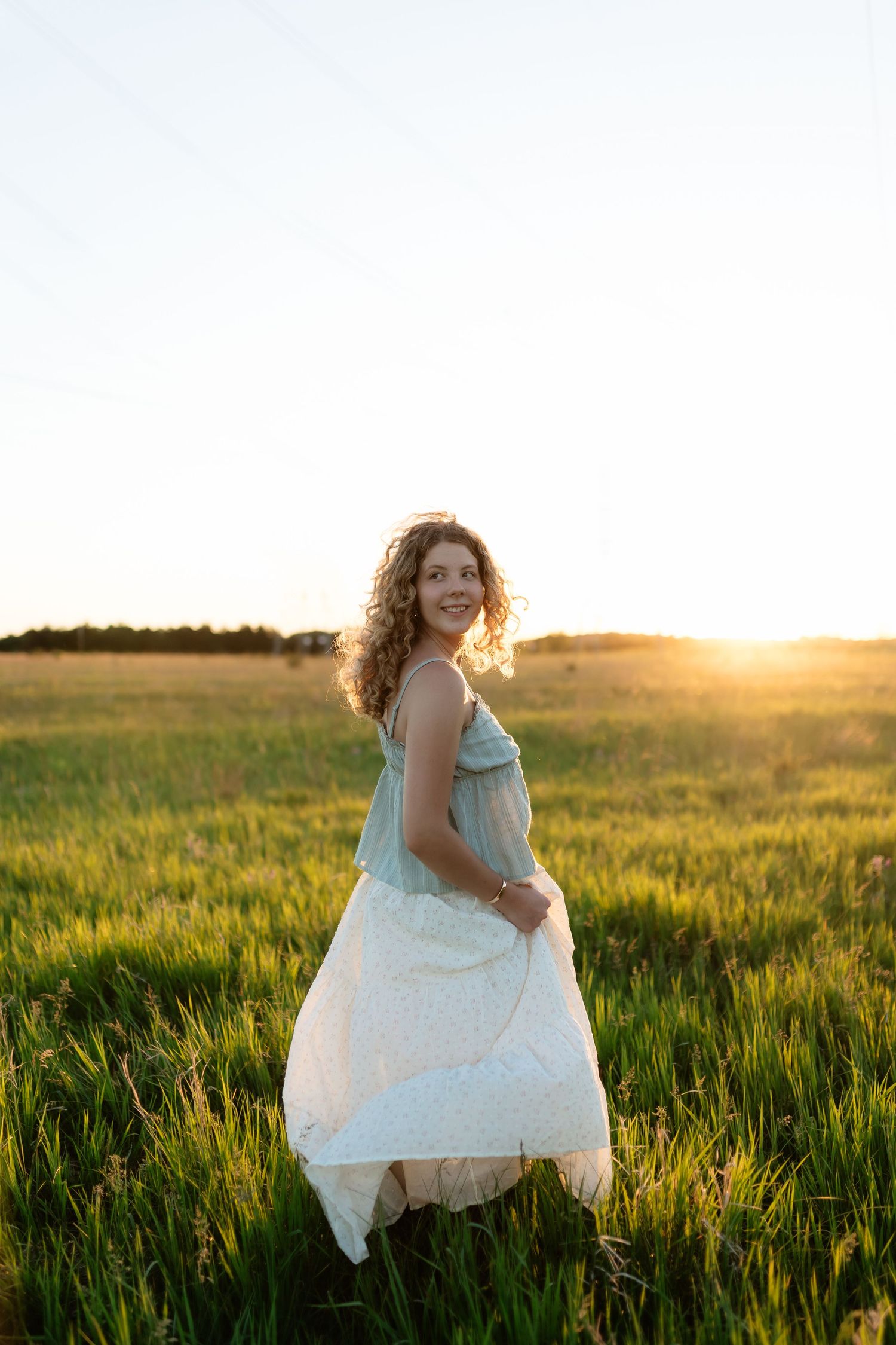 Woman in blue top and white skirt standing in sunlit green field at sunset.