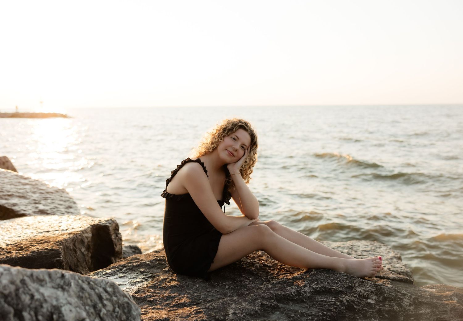 Young woman in black dress sitting on rocky shore at sunset with calm ocean waves in background.
