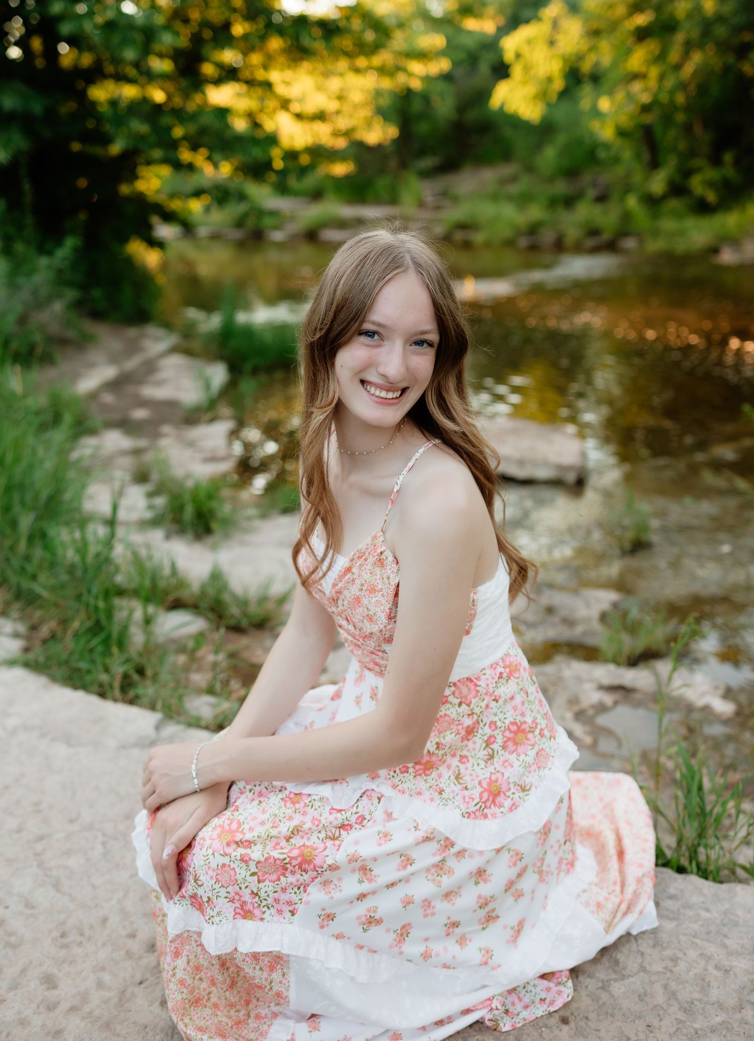 Smiling teenage girl in pink floral dress sits on rocks beside a peaceful wooded creek.