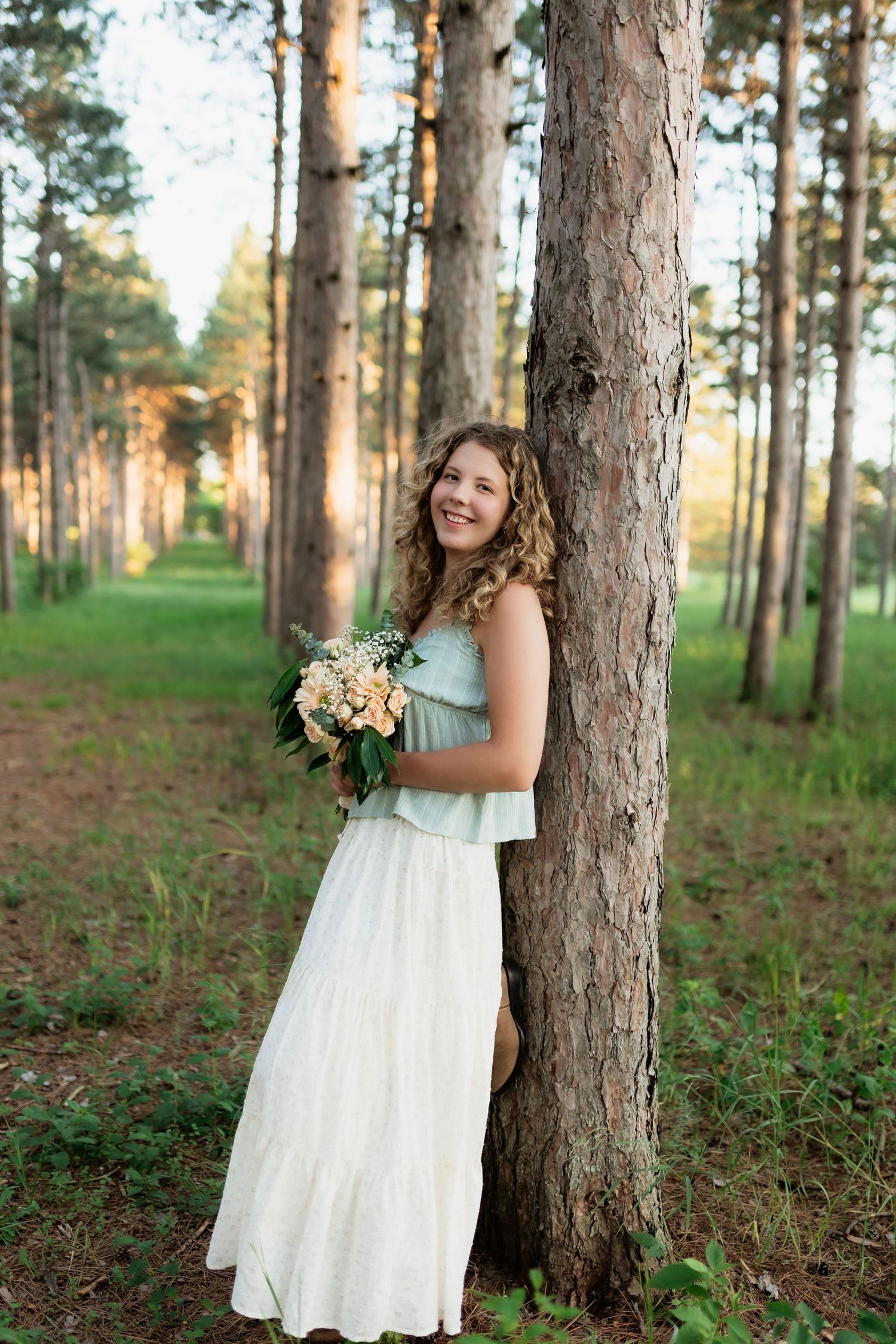 Smiling young woman in mint top and white skirt holding flower bouquet leaning against pine tree in forest.