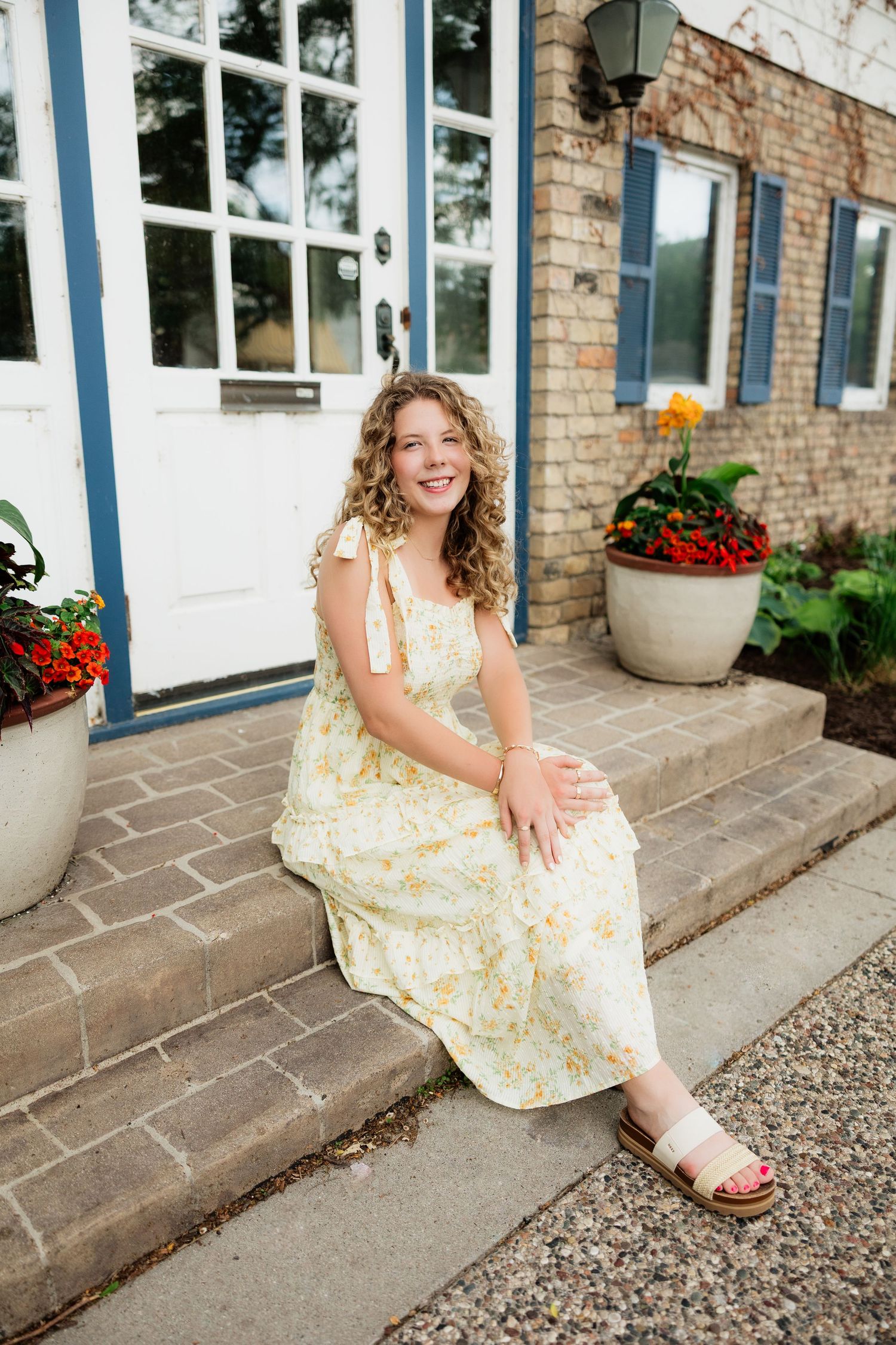 Smiling young woman in yellow floral dress sitting on brick steps outside a charming building with blue shutters.