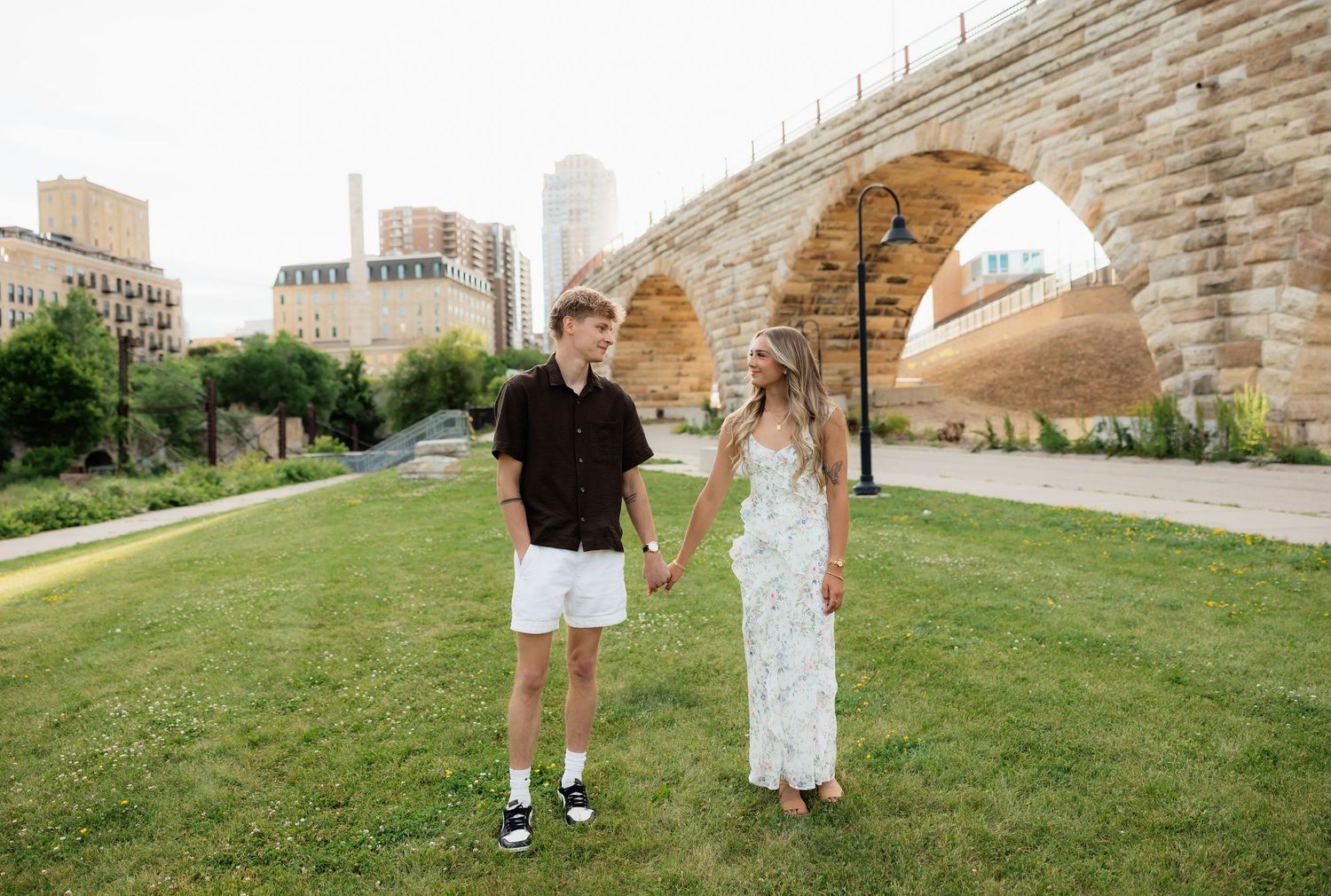 Young couple holding hands while walking on green grass near a historic stone arch bridge in the city.