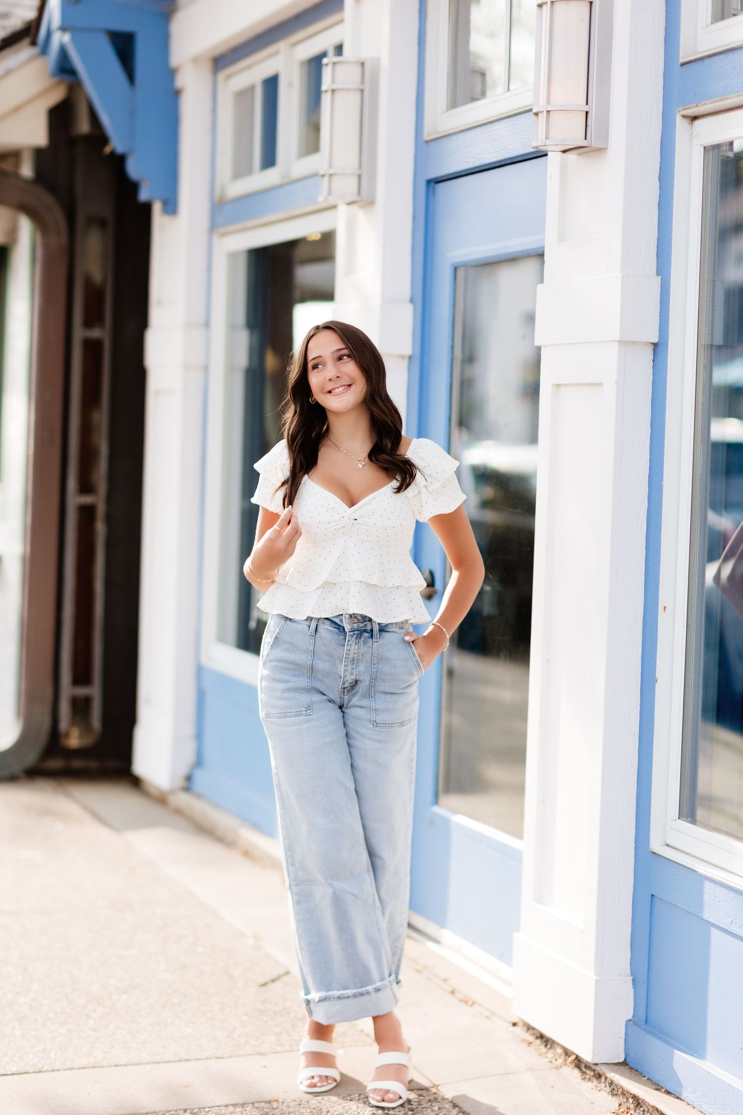 Young woman in white ruffle top and light blue jeans smiling outside a blue and white storefront.