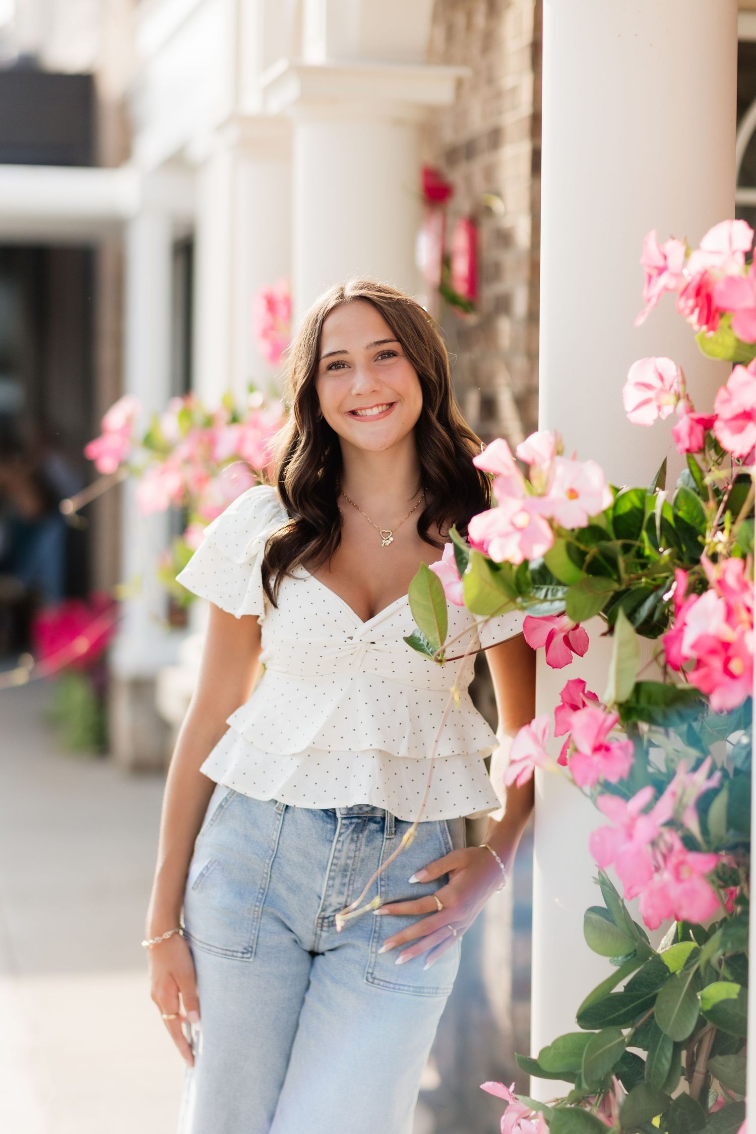 Smiling brunette woman in white ruffled top and jeans posing near pink flowers on a sunny day.