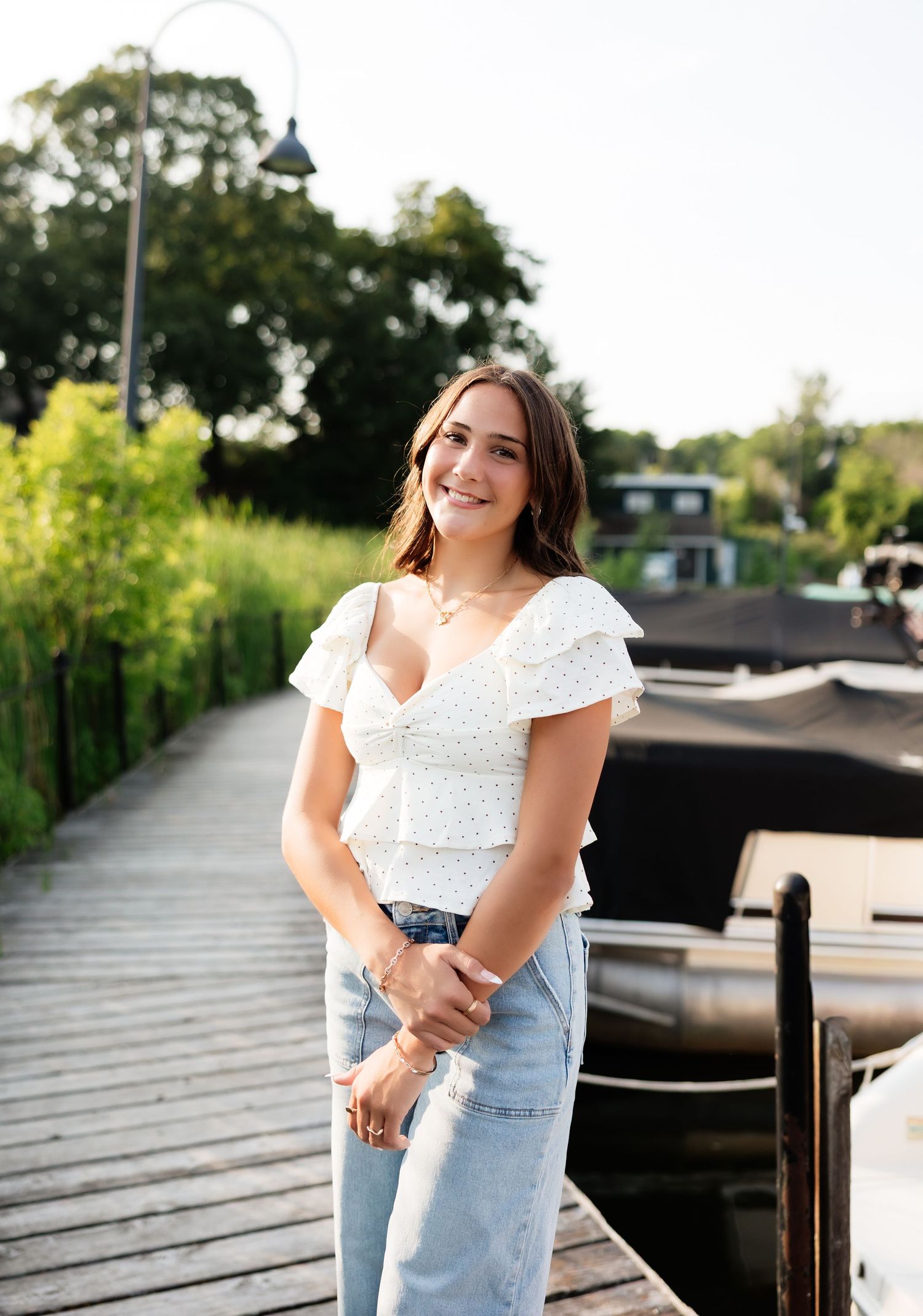 Young brunette woman in white polka-dot crop top and jeans smiles on a sunny wooden marina boardwalk.