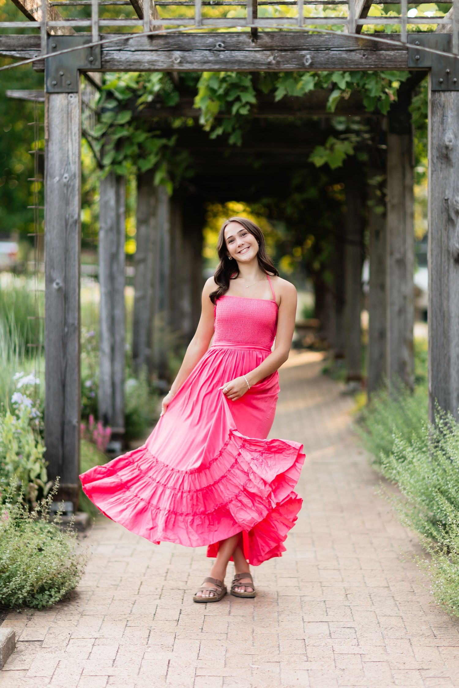 Smiling young woman in pink maxi dress twirling under a wooden pergola in a garden.