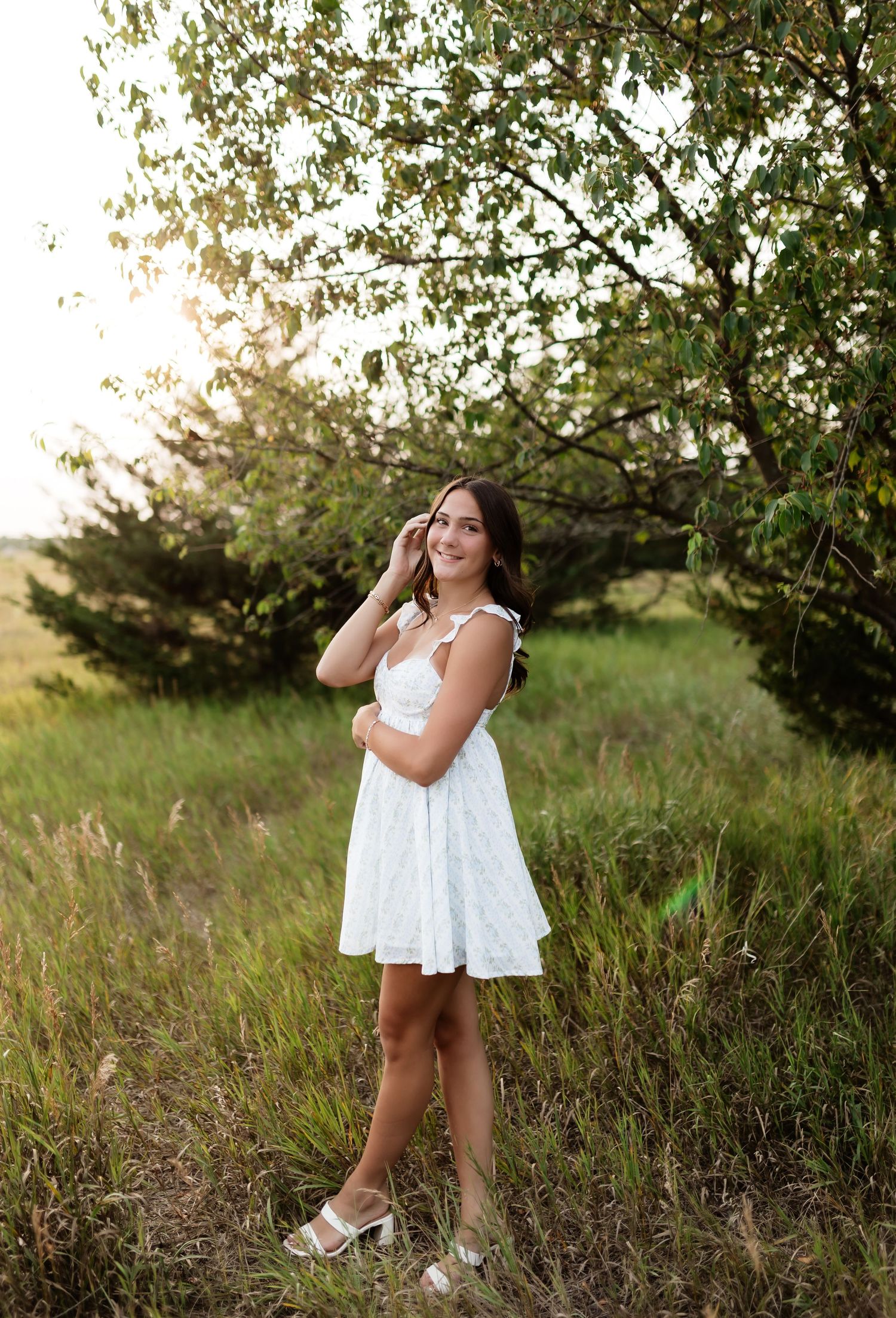 Smiling young woman in a white lace dress stands in a sunlit meadow beside a leafy green tree.
