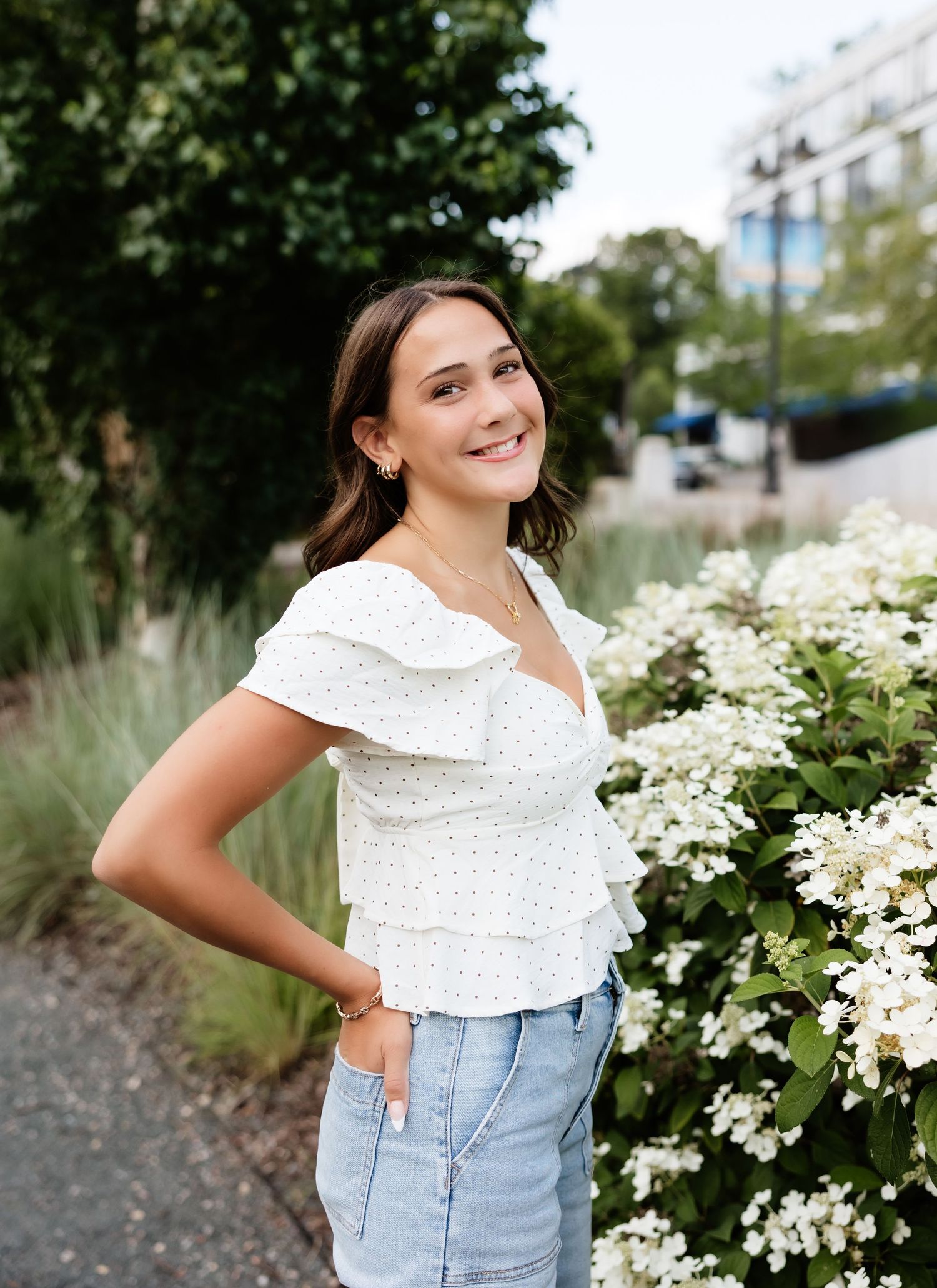 Young woman in white floral top and jeans smiling outdoors near white hydrangea blooms in a garden setting.