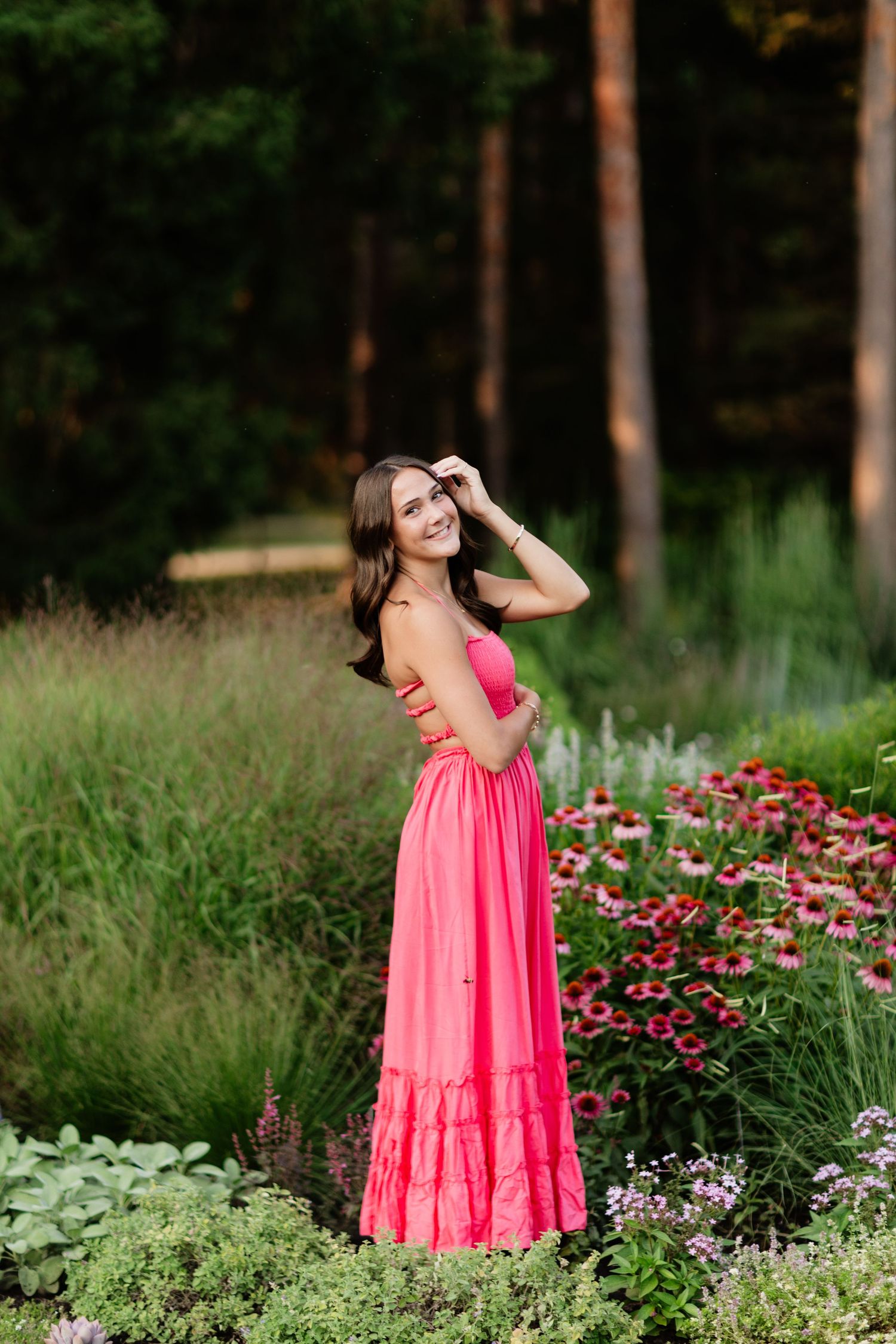 Young woman in pink maxi dress smiling in garden surrounded by colorful wildflowers and tall grasses.