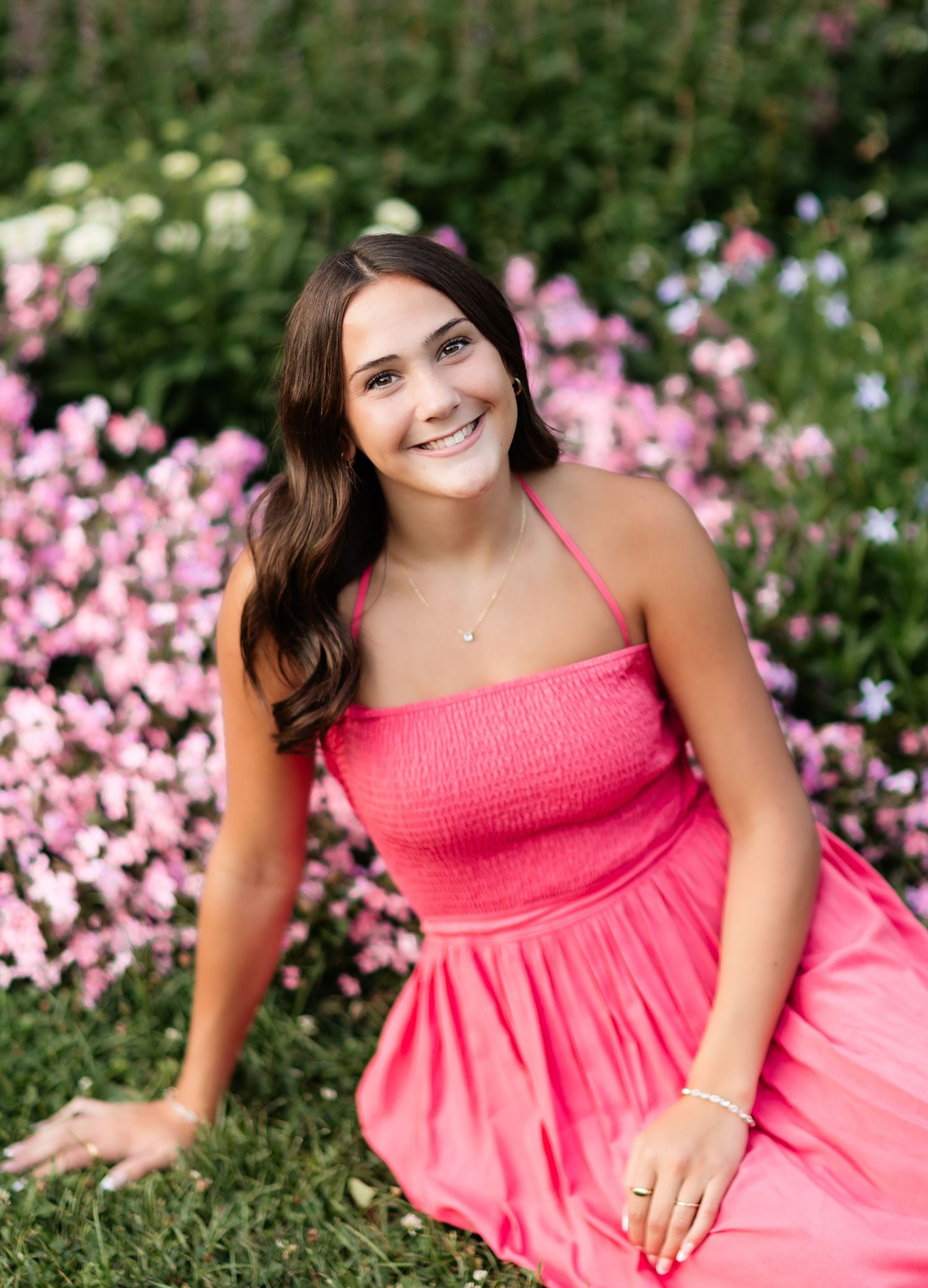 Smiling young woman in a pink halter dress sits among vibrant pink and white flowers in a sunny garden.