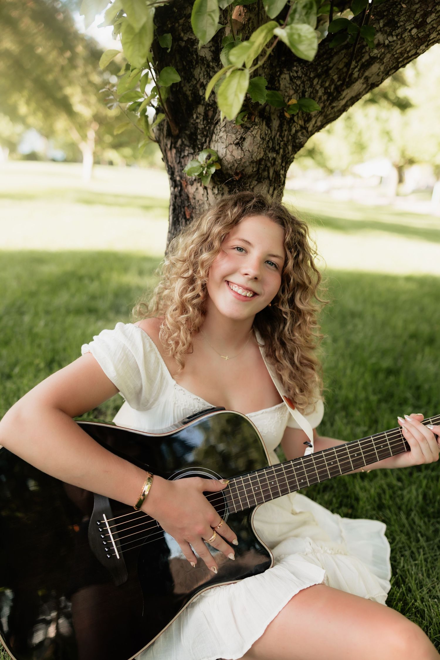 Smiling teenage girl with curly hair playing acoustic guitar under tree in green park.