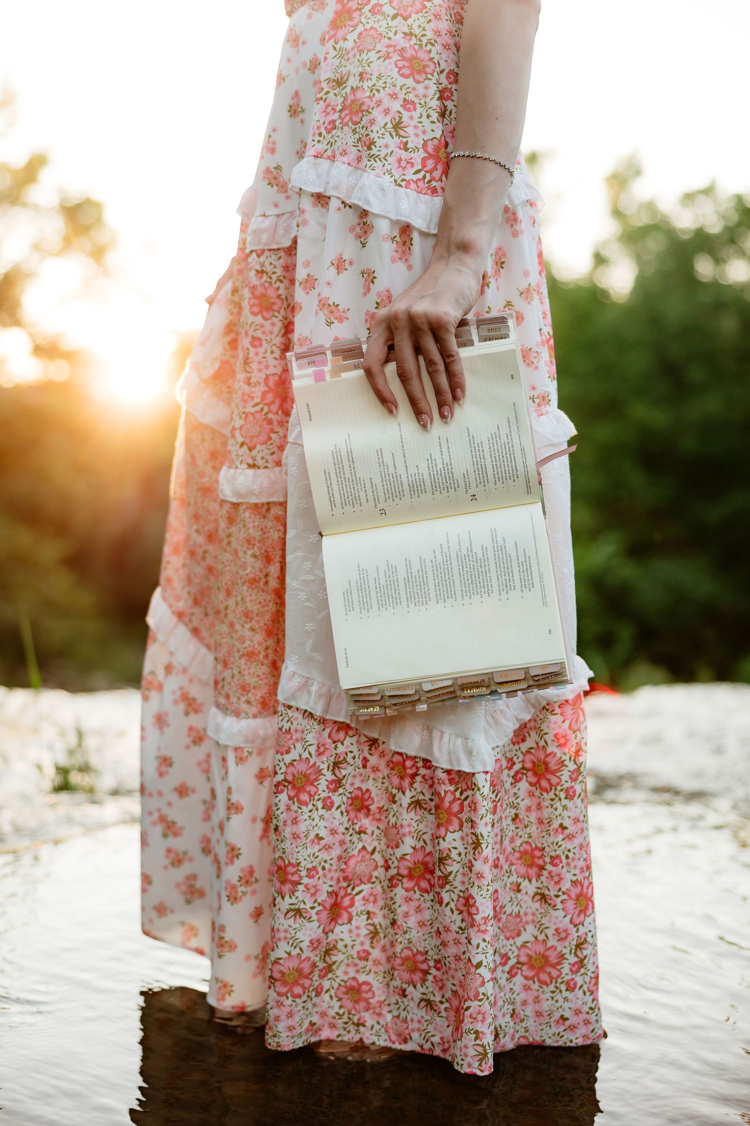 Close-up of woman in floral dress holding open book at golden sunset by a creek.