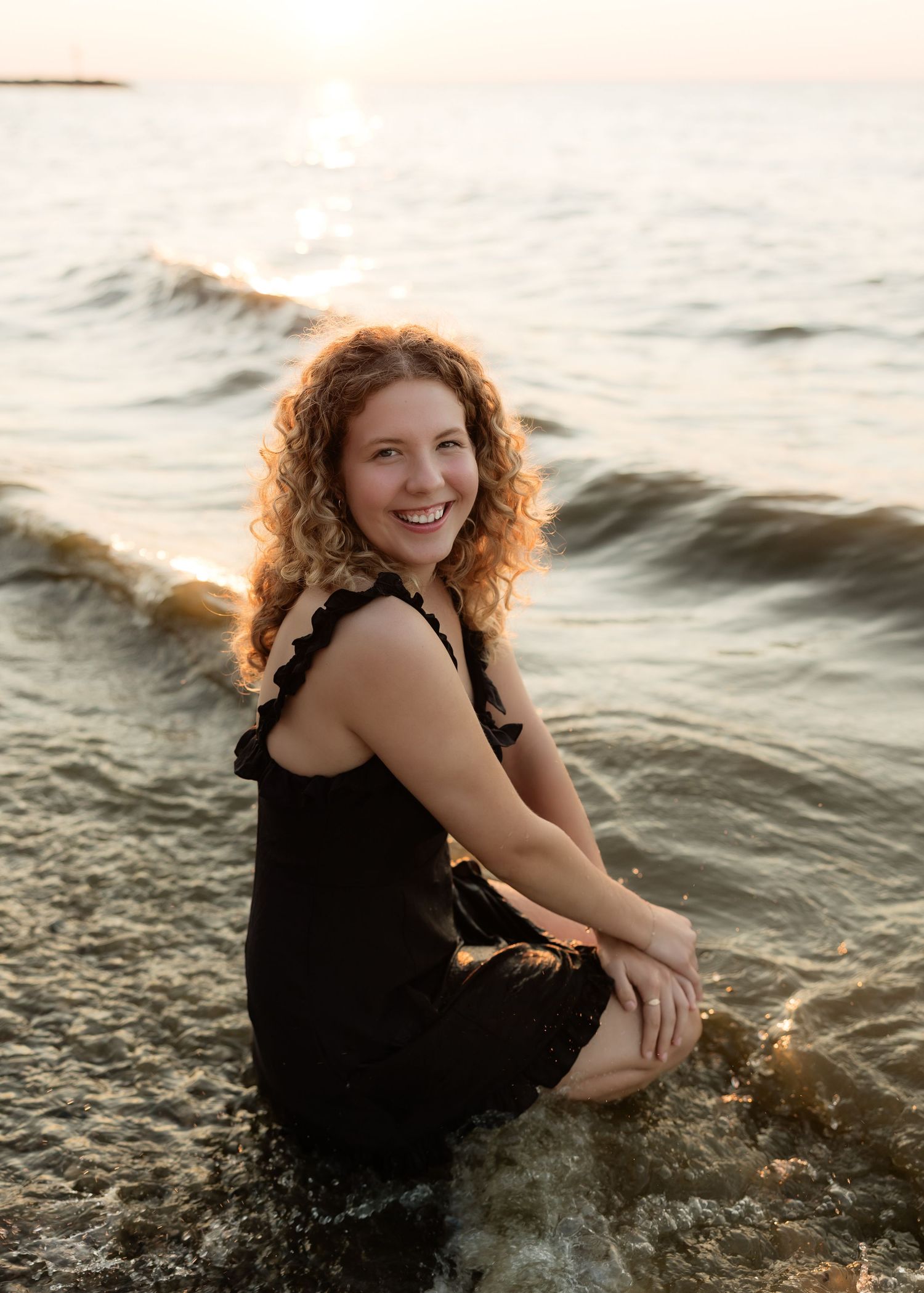 Young woman with curly red hair sits smiling in ocean waves at sunset, wearing a black dress.
