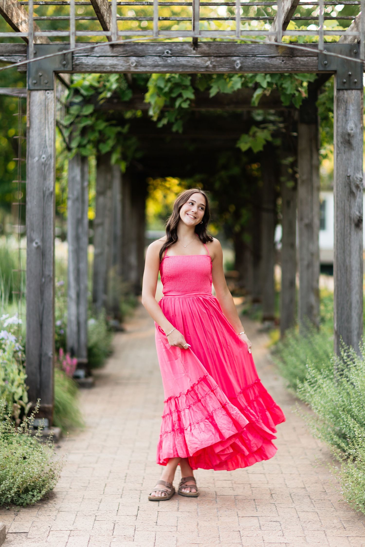 Young woman in bright pink strapless maxi dress twirling under a wooden garden pergola archway.