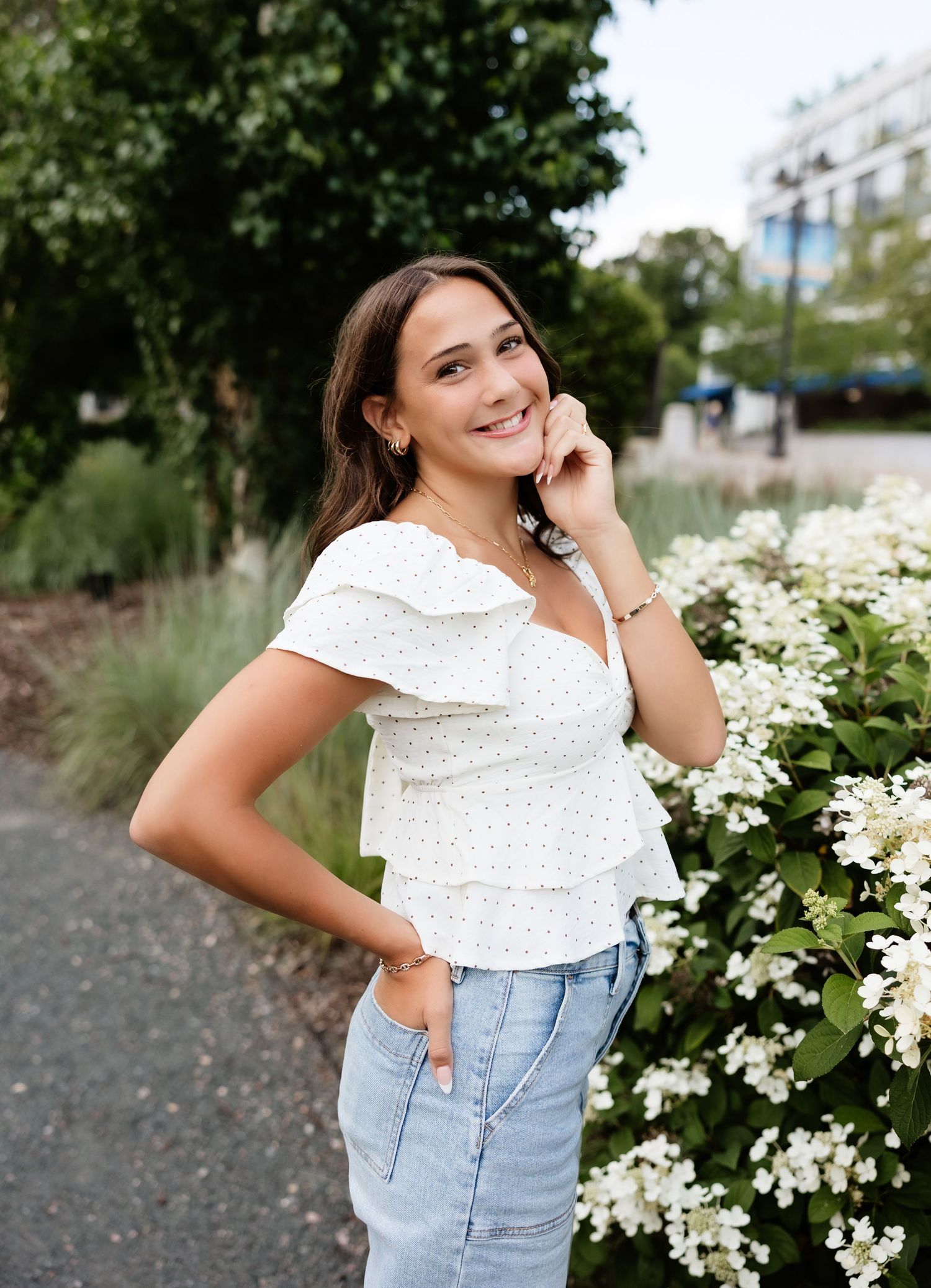 Young woman in white ruffled crop top and jeans smiling beside white hydrangea blooms outdoors.