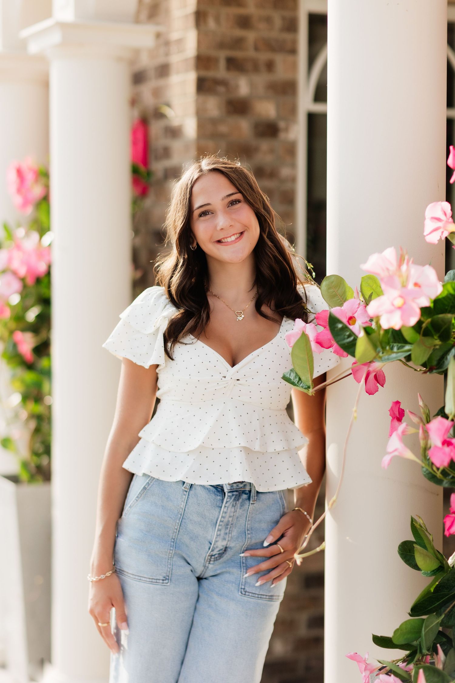 Smiling young woman in white eyelet top and jeans posing between white columns with pink flowers.