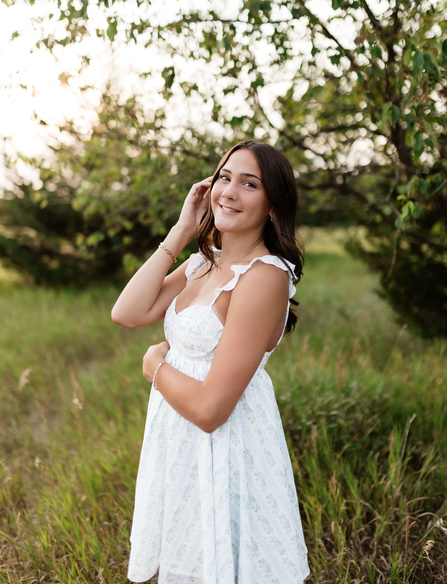 Smiling brunette woman in white floral dress posing in golden hour sunlight among green trees.