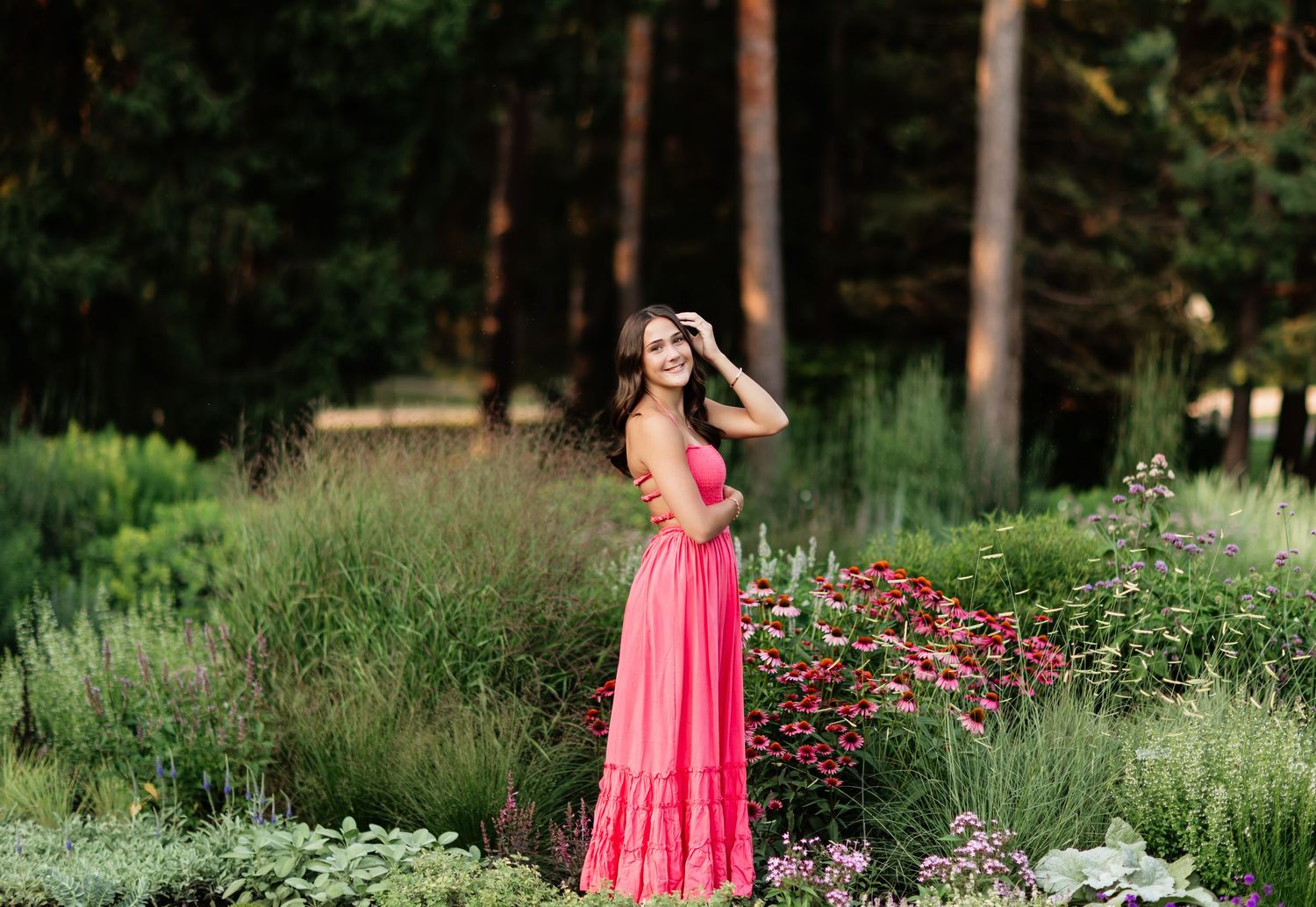 Young woman in pink maxi dress standing among colorful garden flowers near tall pine trees.