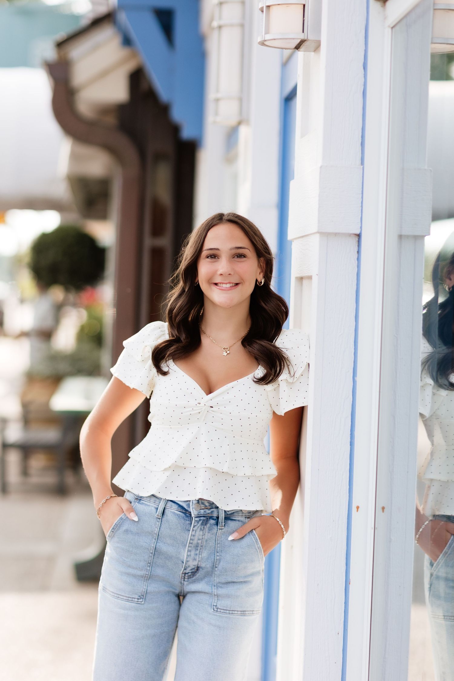 Brunette woman in white ruffled top and light blue jeans leaning against a blue and white building exterior.